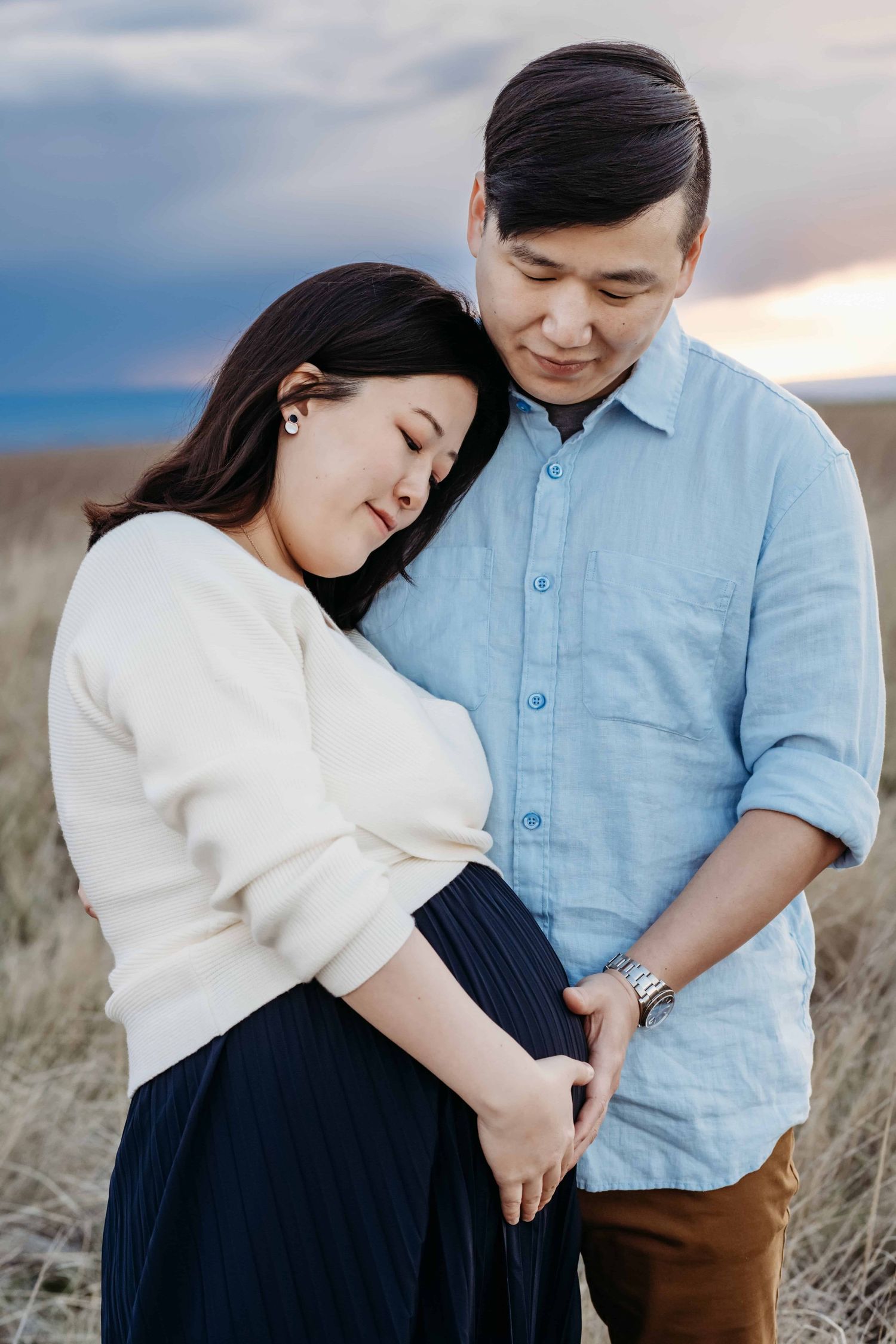 A couple expecting a baby looking down at the pregnant belly with a blue sunset in the background.