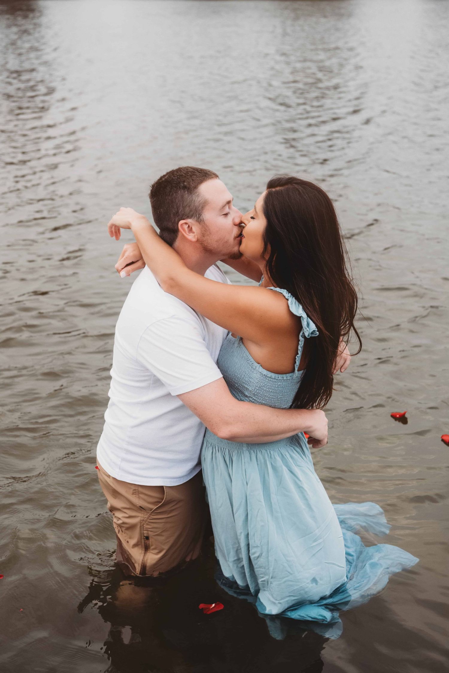 A couple standing in a river and kissing with red rose petals surrounding them.