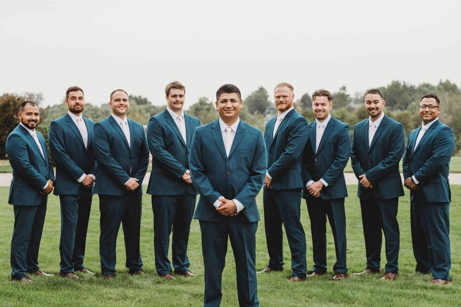 A groom standing with his eight groomsmen standing behind him while they all smile at the camera.