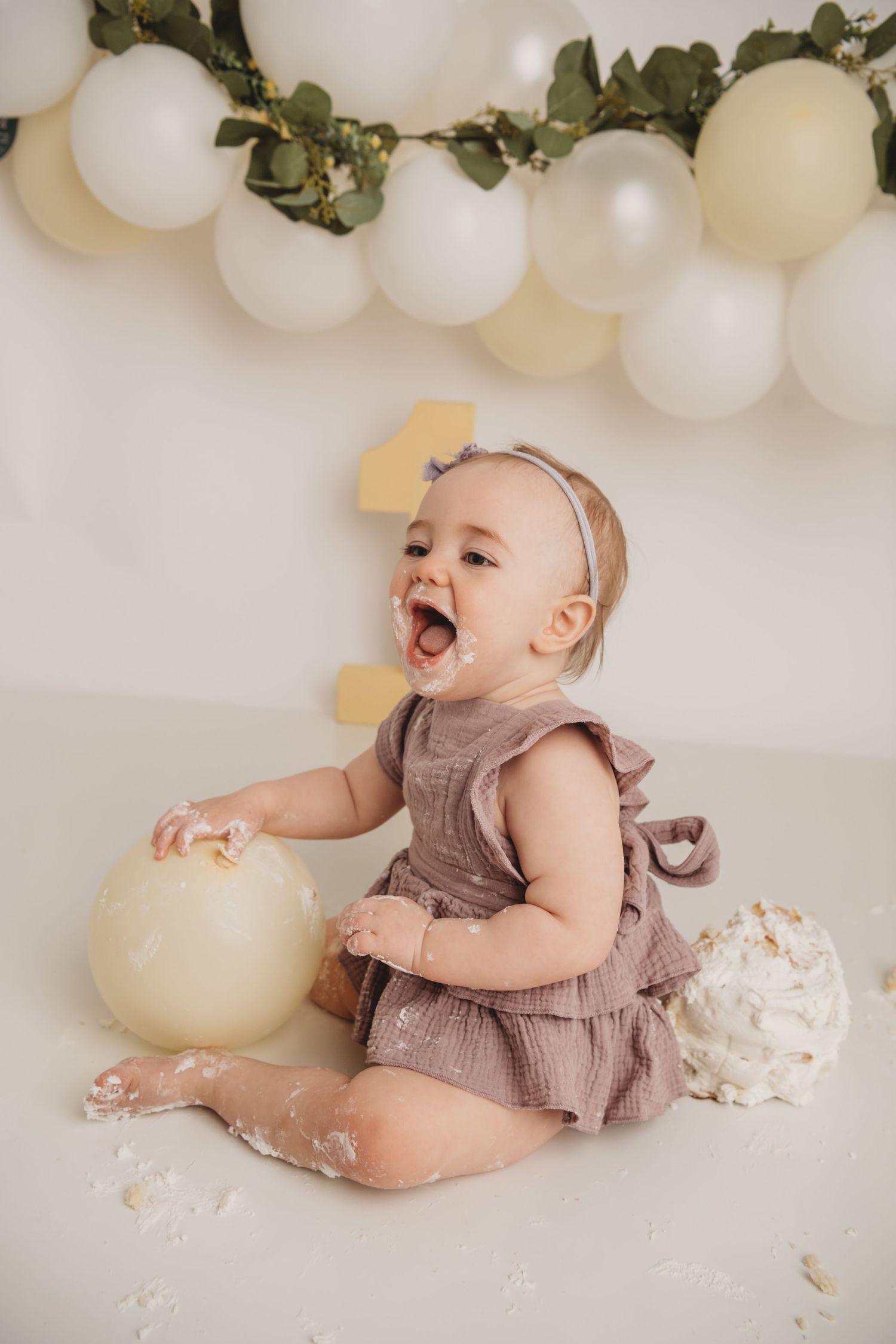 A baby wearing a purple romper covered in cake with a balloon garland in the background.