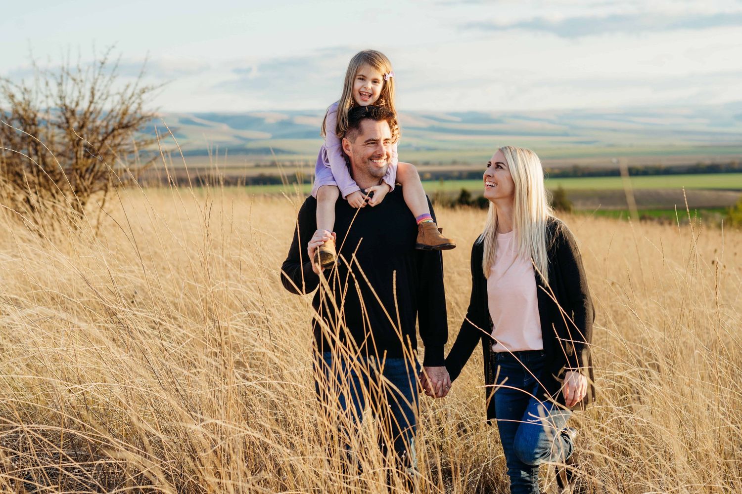 A candid portrait of a family of three with a little girl on her dad's shoulders walking through a field with green and blue hills.