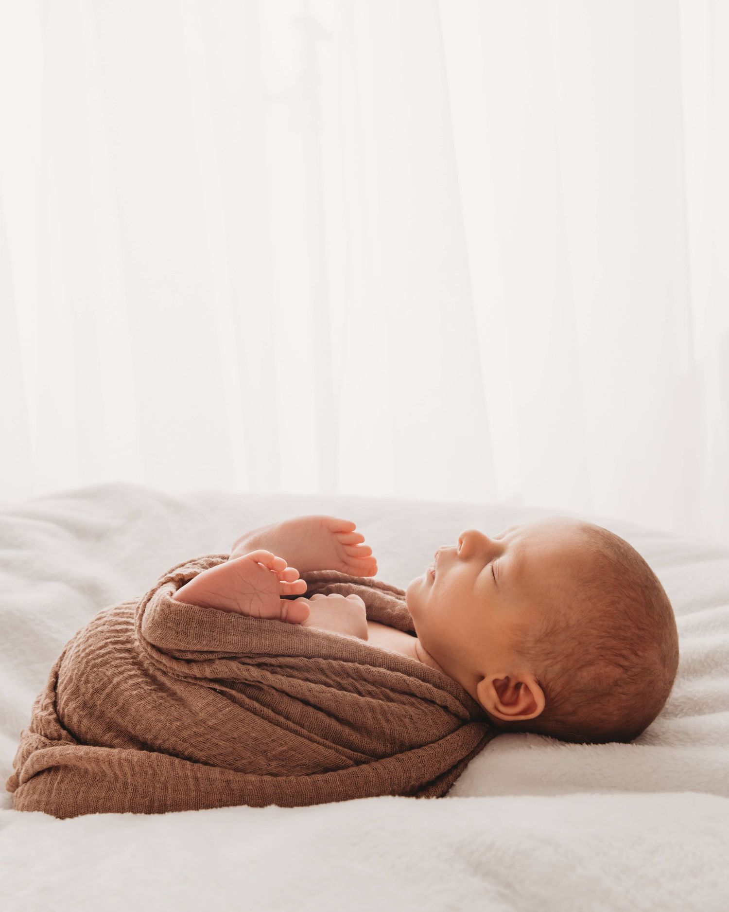 A backlit photo of a newborn wrapped in brown sleeping on a white beanbag.