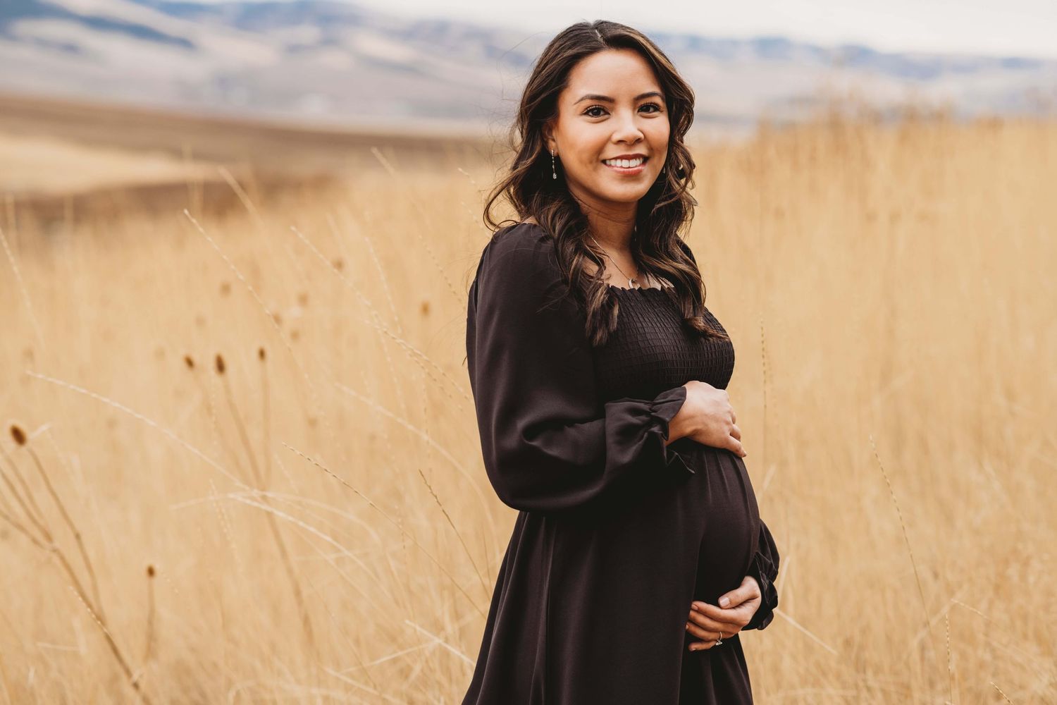 A pregnant woman wearing a black dress holds her baby bump while smiling at the camera with rolling hills in the background in Walla Walla.