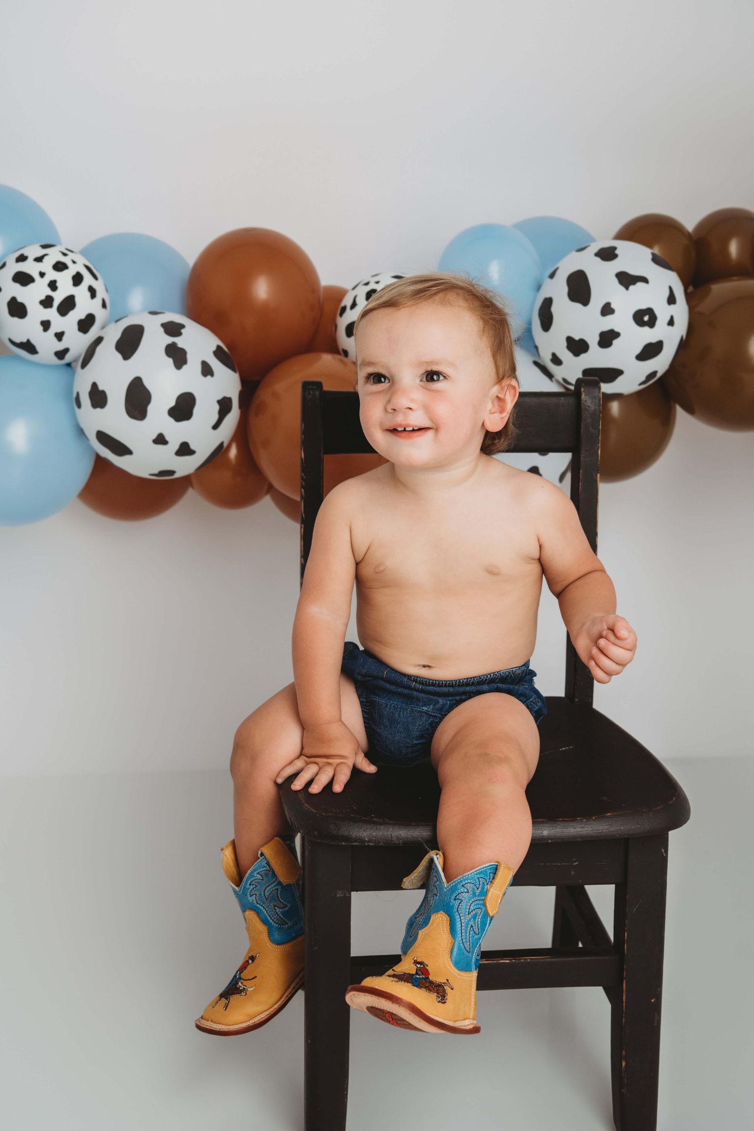 Baby milestone portrait by a Walla Walla photographer featuring a smiling toddler sitting on a chair with western boots.