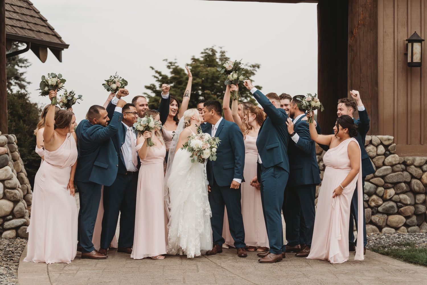 A large wedding party dressed in pink and navy surrounding the bride and groom as their share a kiss before their wedding ceremony.