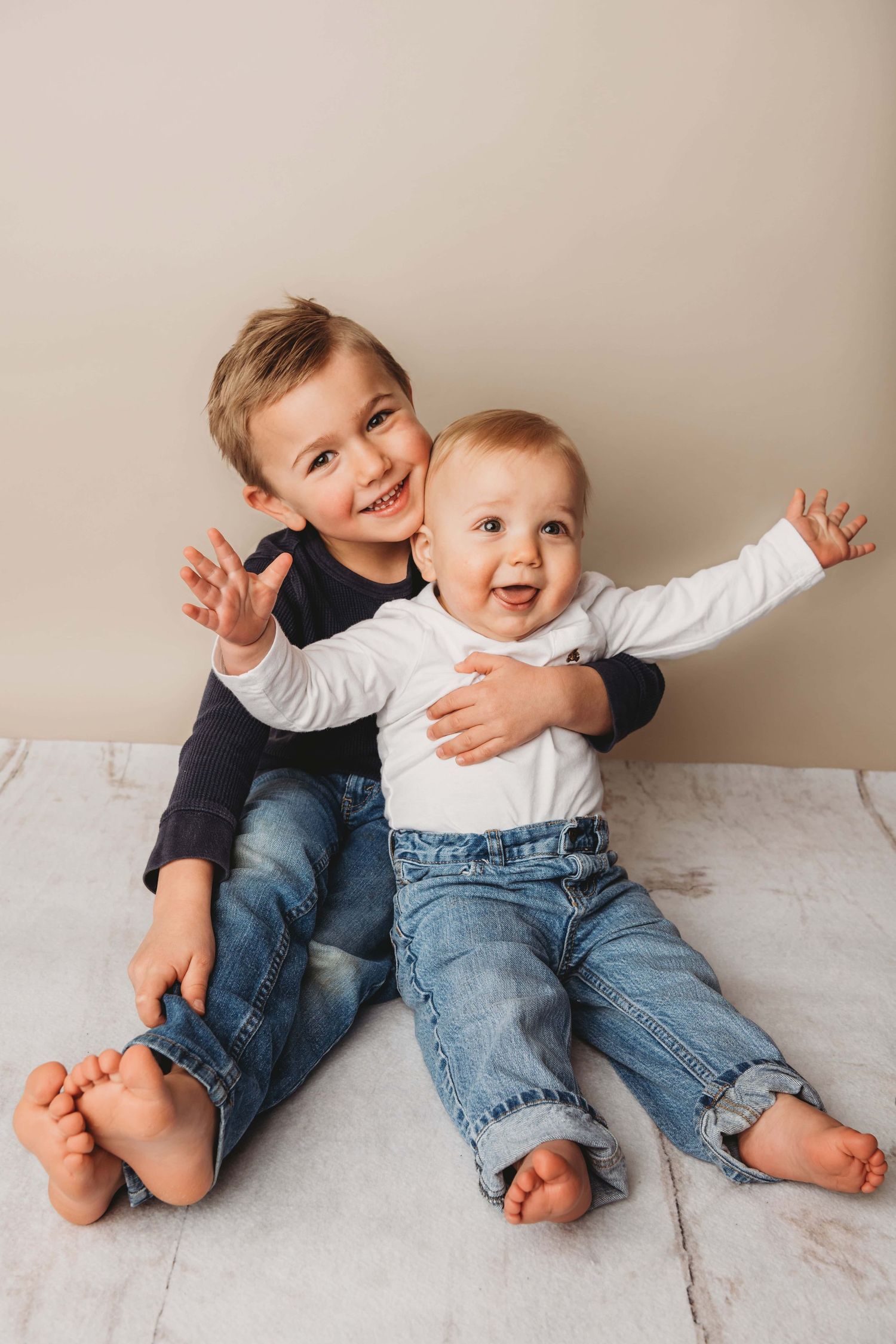 Two children sitting on a white wooden floor while they hug and smile.