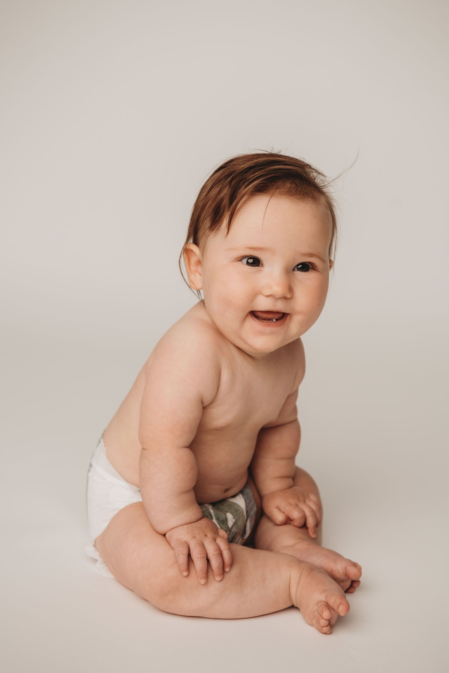 A portrait of a baby girl wearing a diaper sitting up and smiling on a white backdrop.