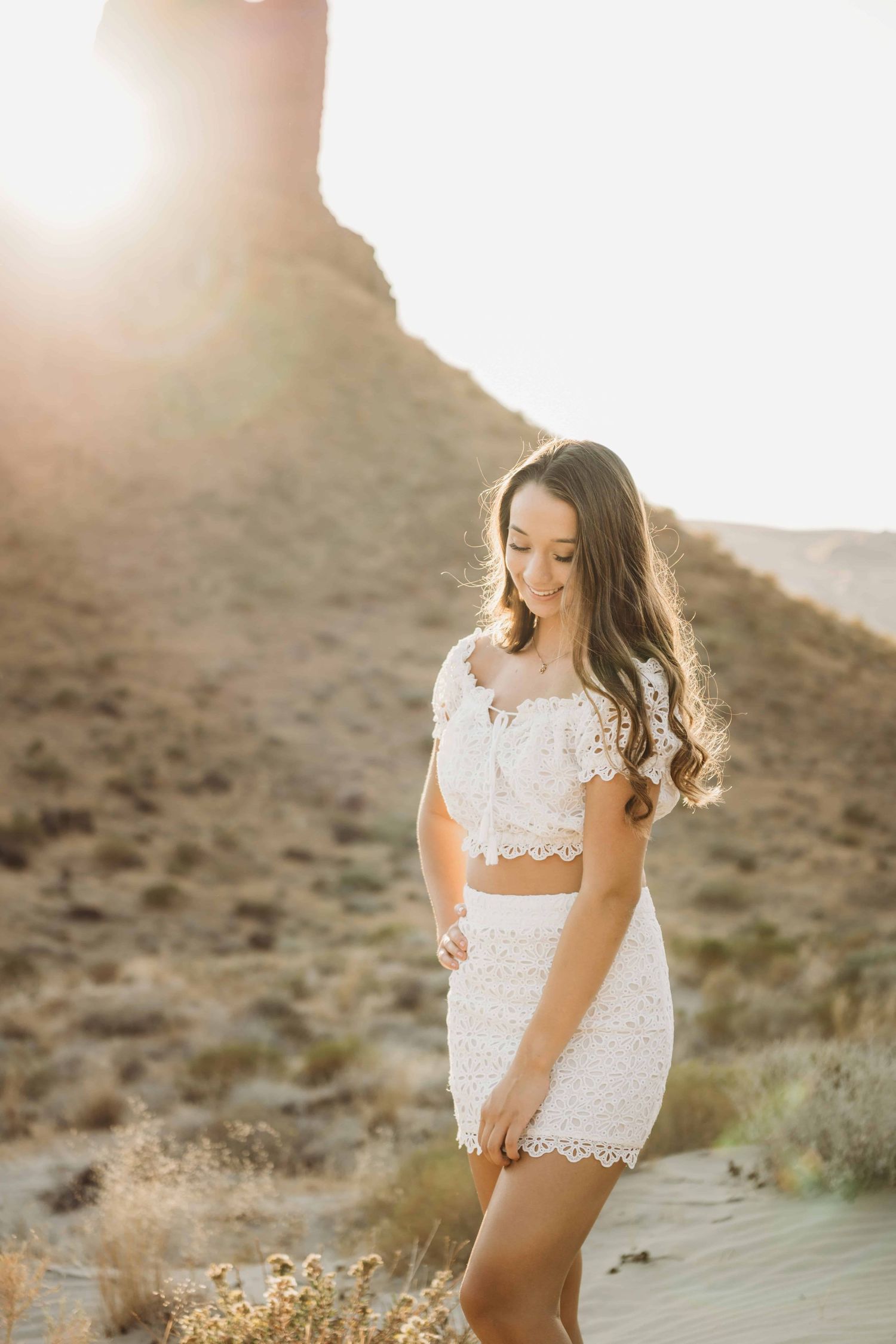 A senior wearing all white standing in a sandy area with a rock formation in the background and the sunlight glowing behind her.