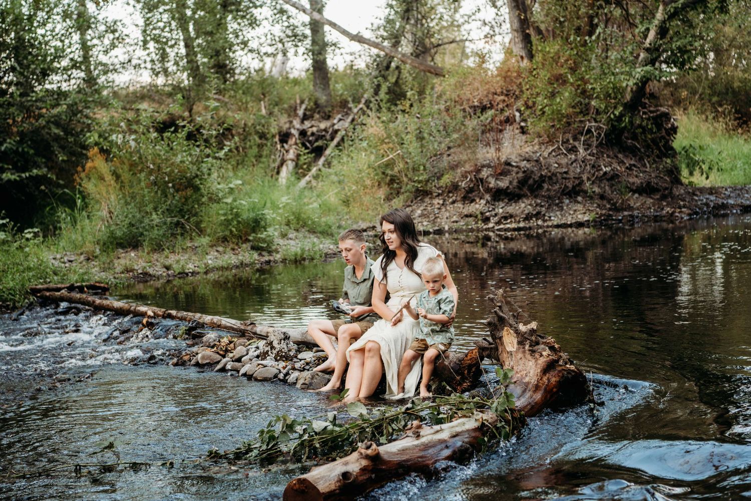 A photographer captures a portrait of a mom and her two sons sitting on a log in a creek in Walla Walla.