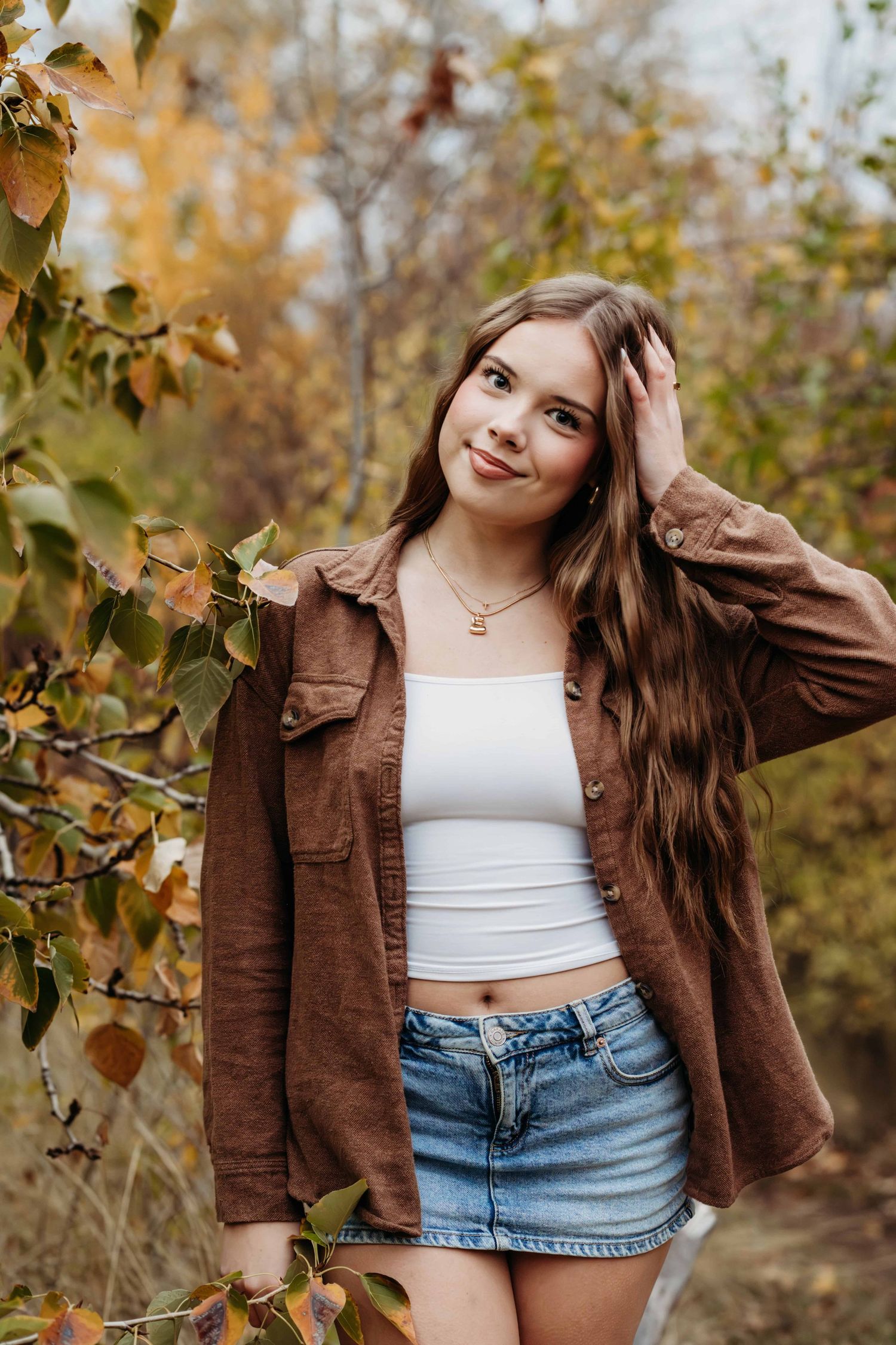 A young woman looking at the camera and smiling with her hand on her head, surrounded by fall foliage.