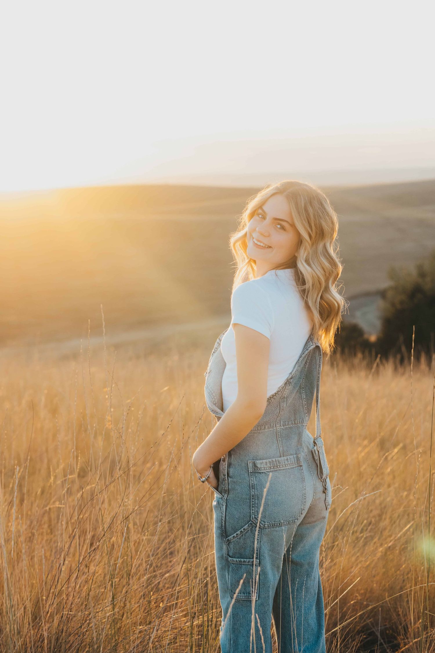 A portrait of a senior wearing overalls looking over her shoulder and smiling during golden hour in Walla Walla.