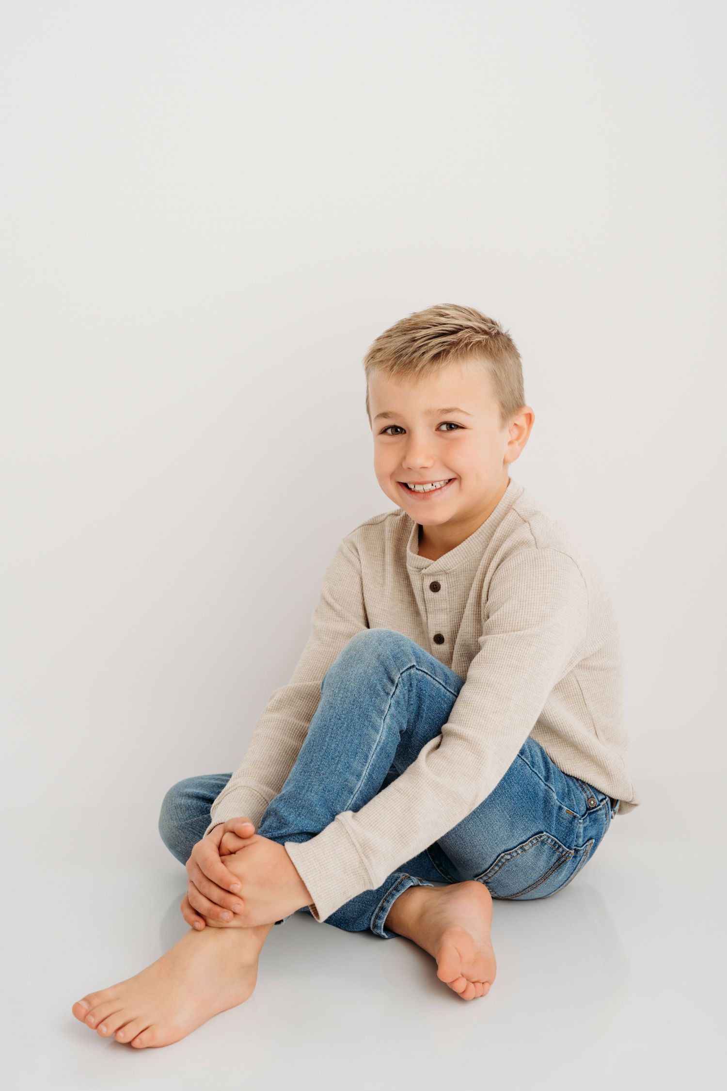 A portrait of a young boy sitting on a white backdrop with his legs crossed.