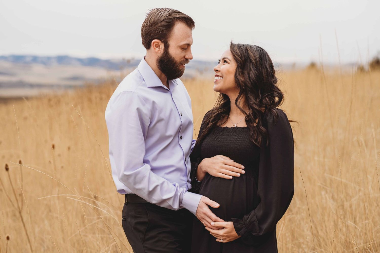 An expecting couple looks at each other, cradling the baby bump while standing in a field.