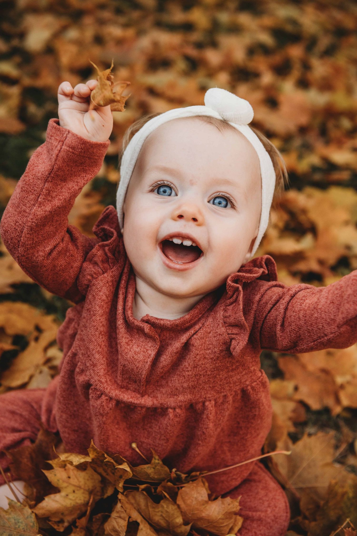 A baby wearing fall tones sitting in a pile of leaves and laughing.