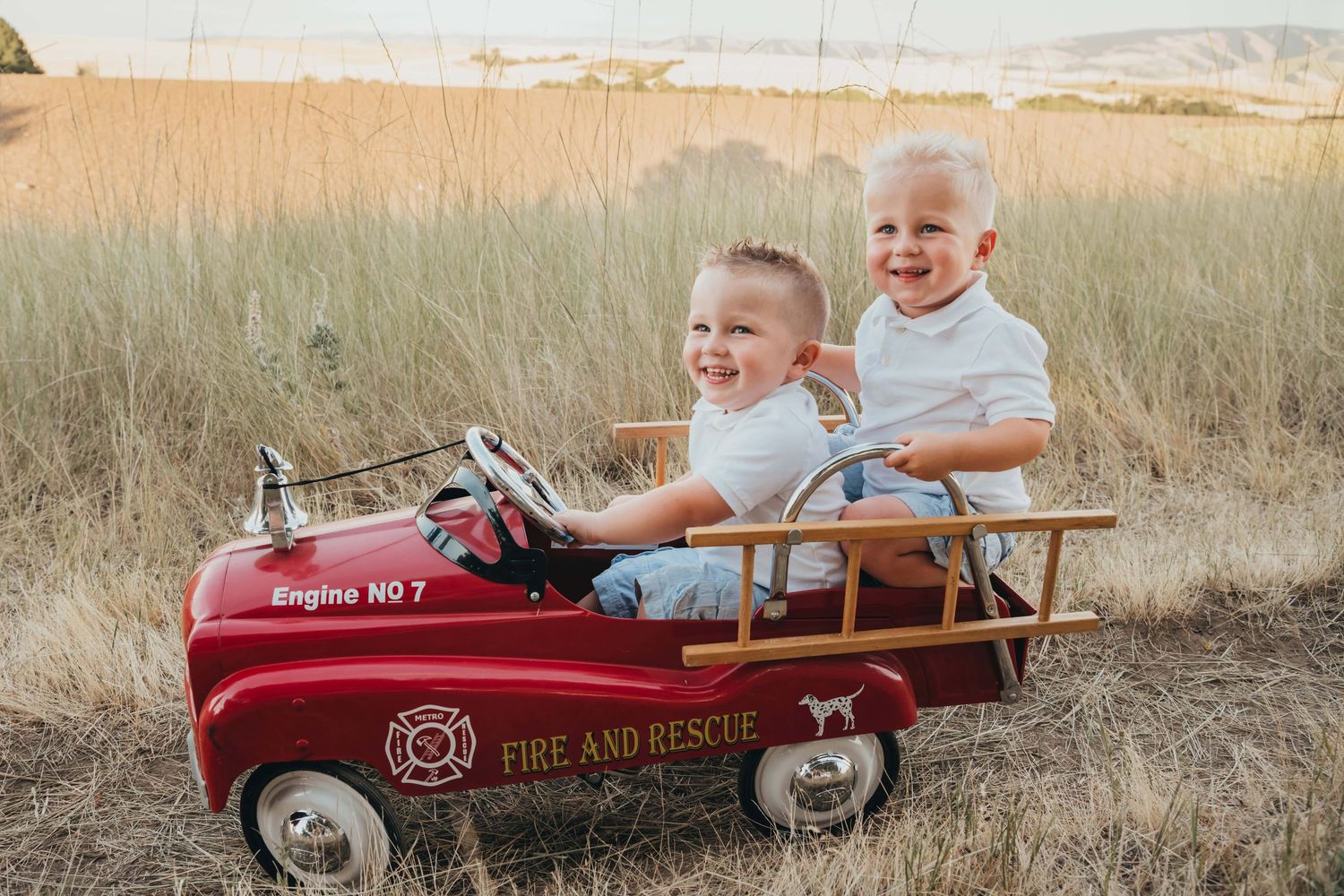 Two toddler boys sitting in a firetruck on a dirt path.