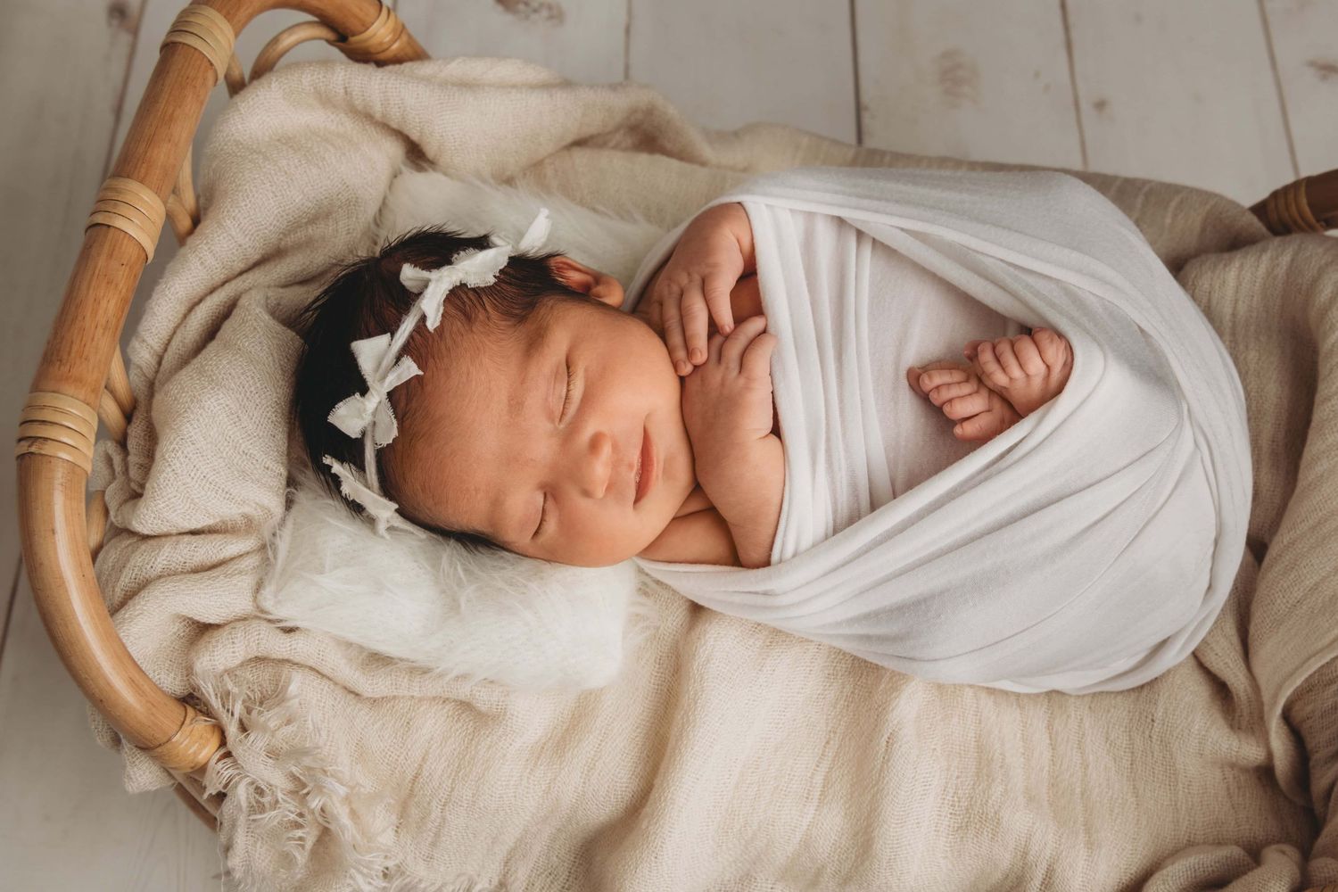 A newborn wrapped in white with a white headband sleeping on a brown bed.