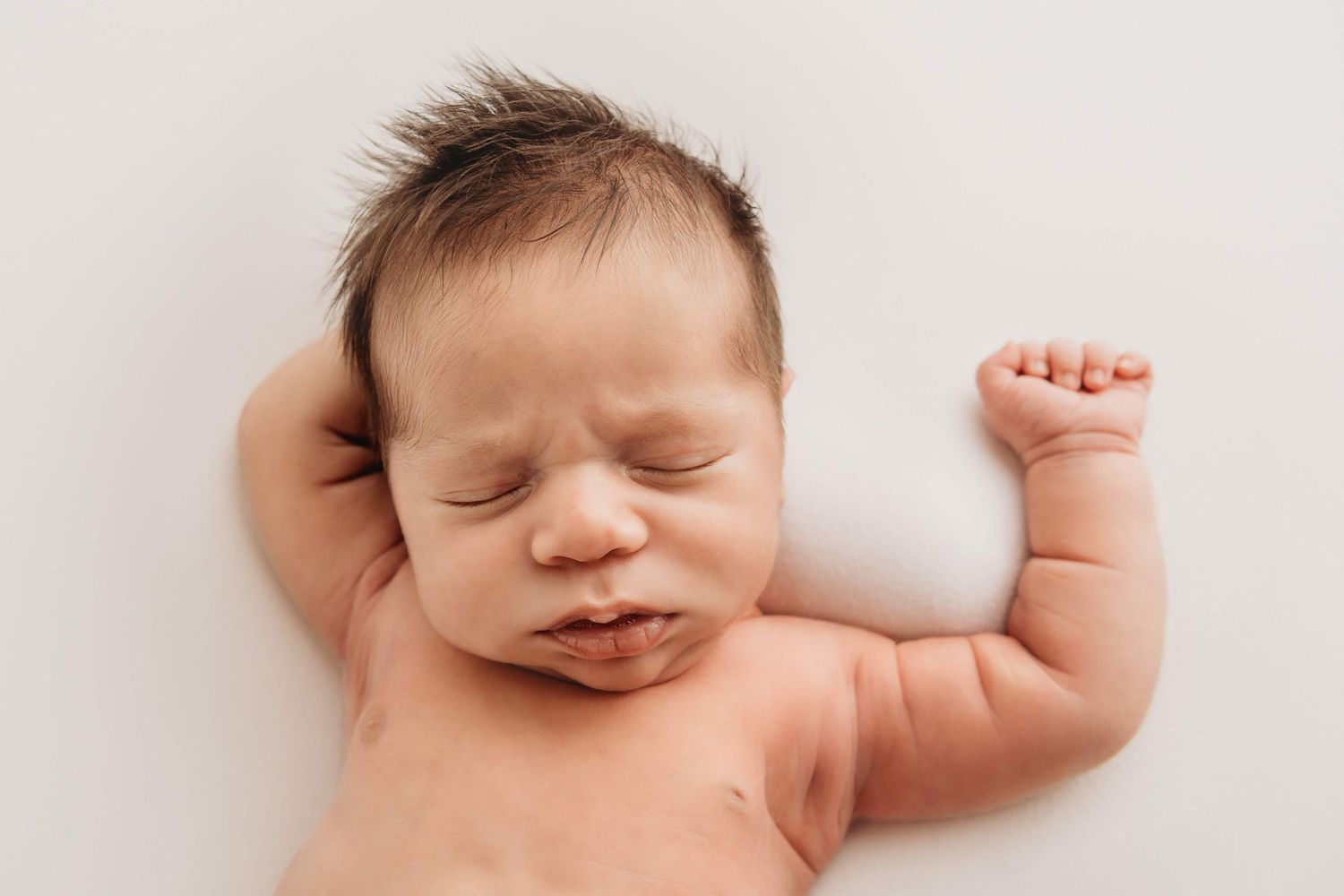 A newborn sleeping with his arms up on a white blanket.