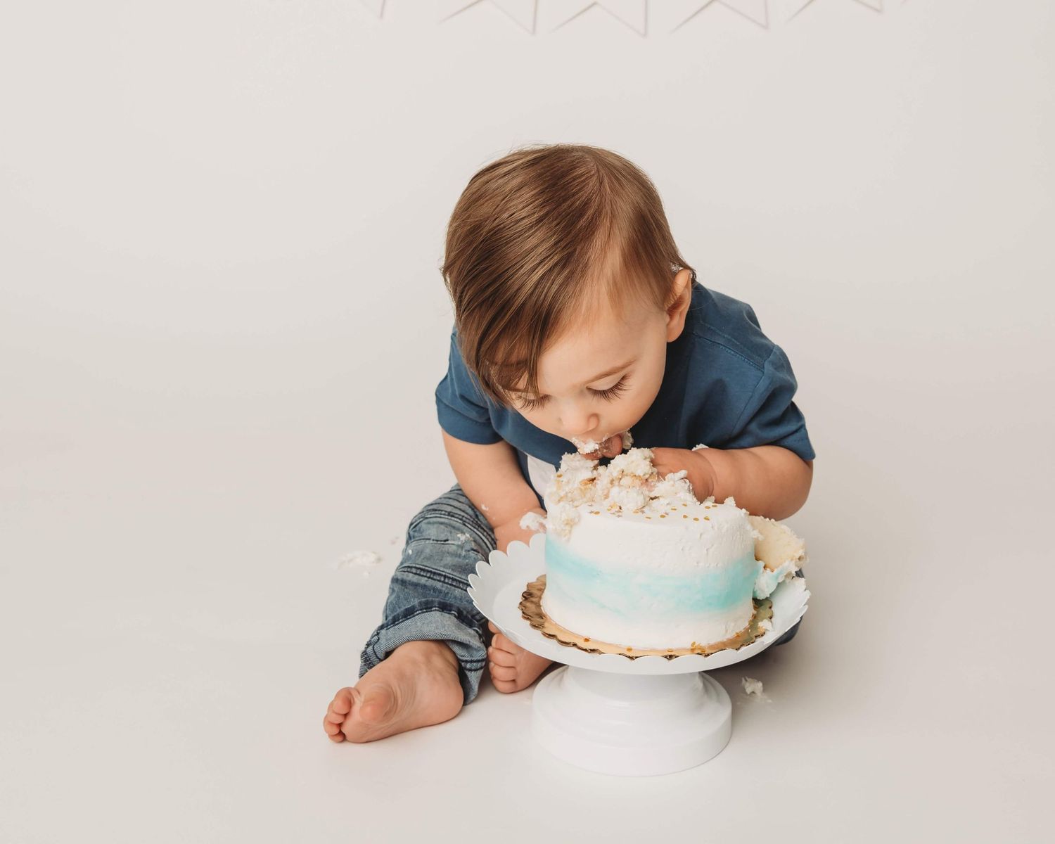 A one year old baby puts his face into a white and blue cake on a white cake stand.
