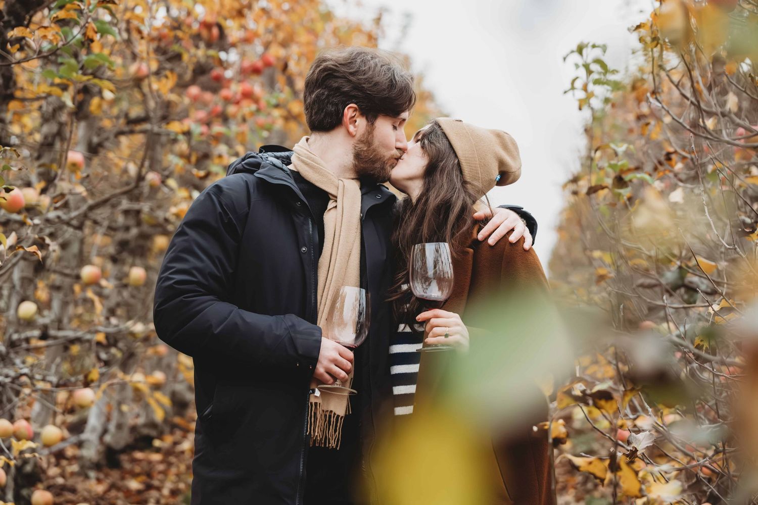 A couple holding wine glasses and kissing in between apple orchard trees.