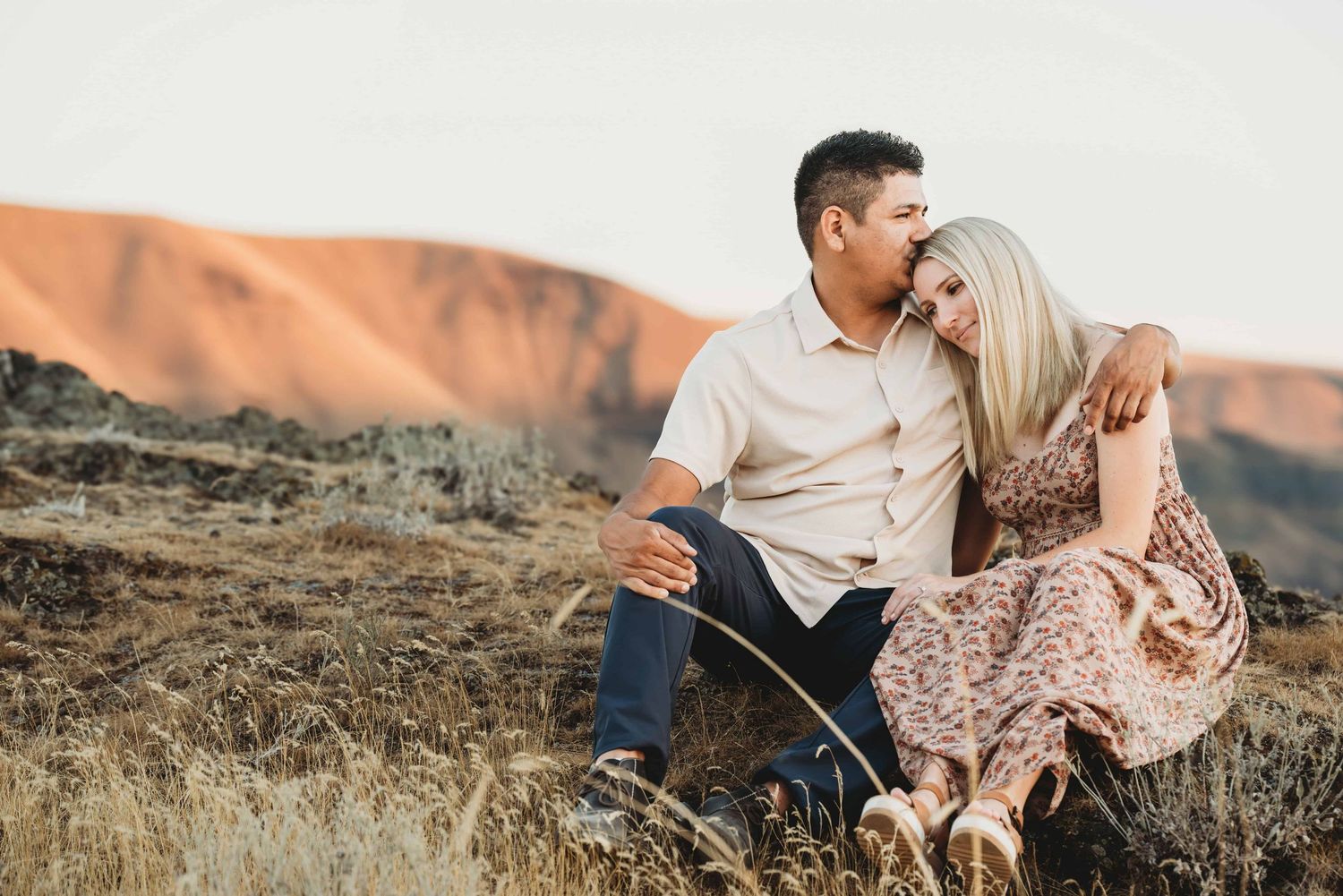 A couple snuggling on top of a mountain while the husband kisses his wife's head.