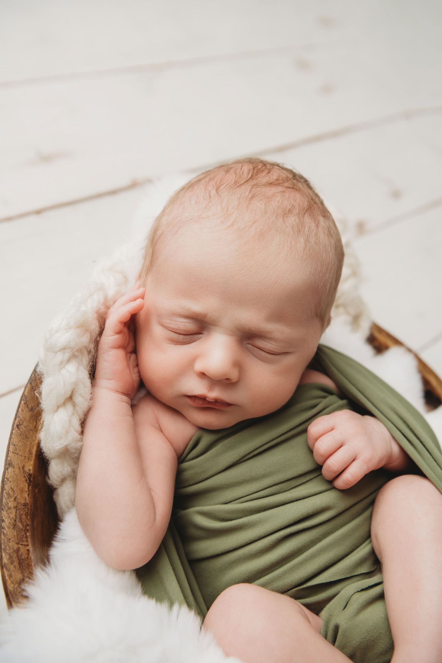 A newborn boy wrapped in olive green sleeping in a wooden bowl on neutral toned fabrics.