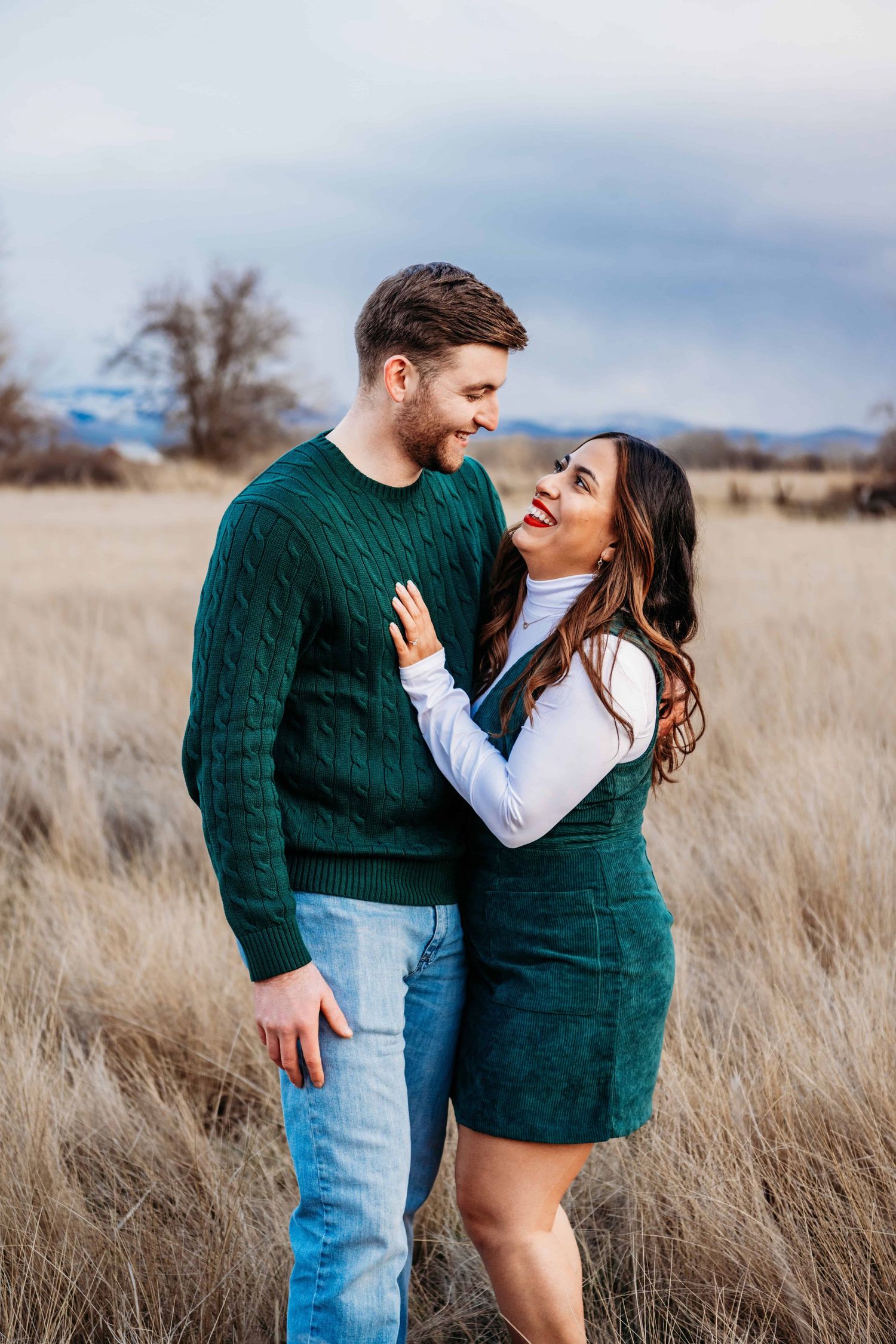 A couple wearing deep green clothing looks at each other smiling while standing in a field with a purple sunset.