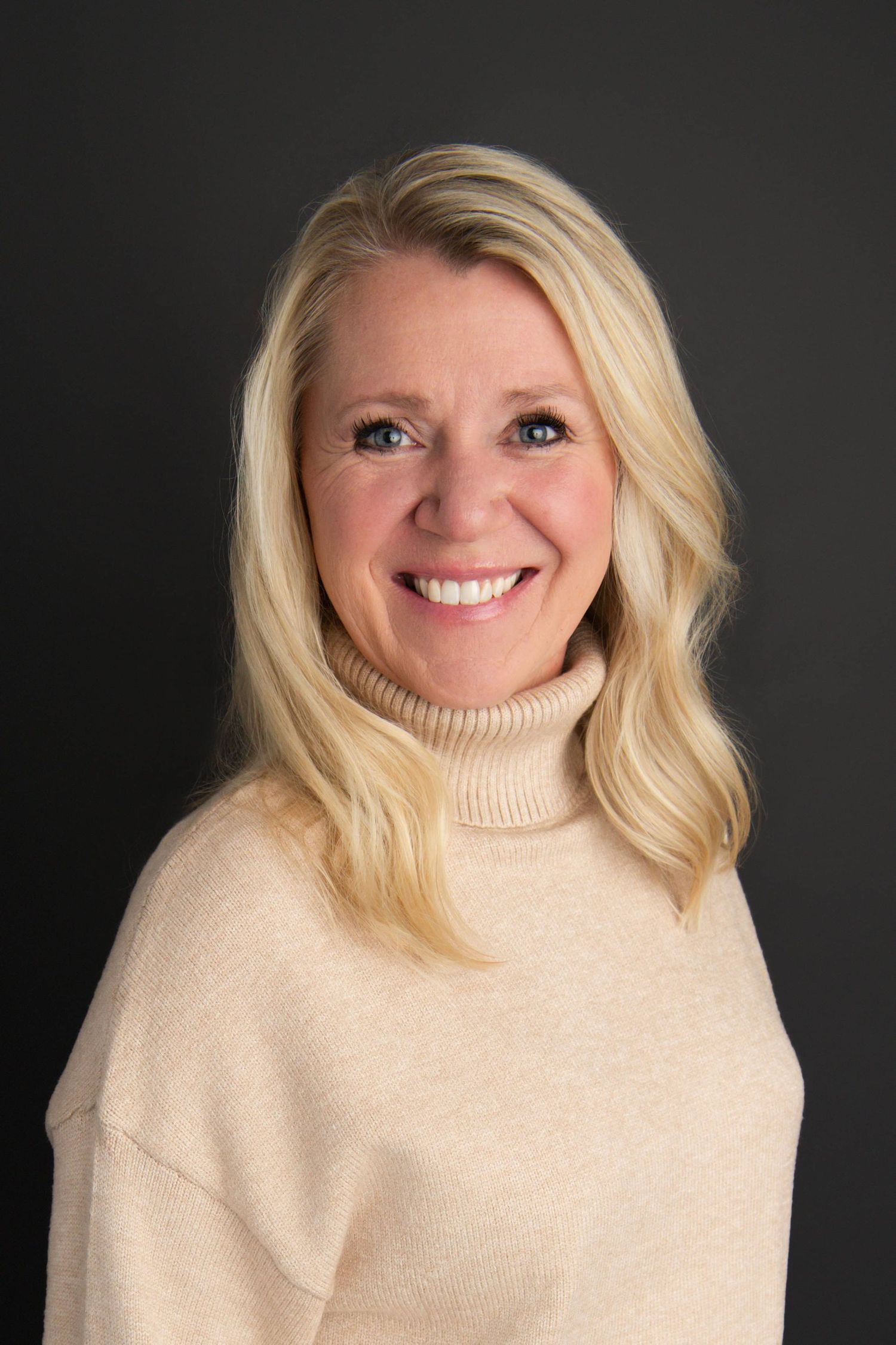 A headshot of a woman in a tan shirt on a black backdrop.