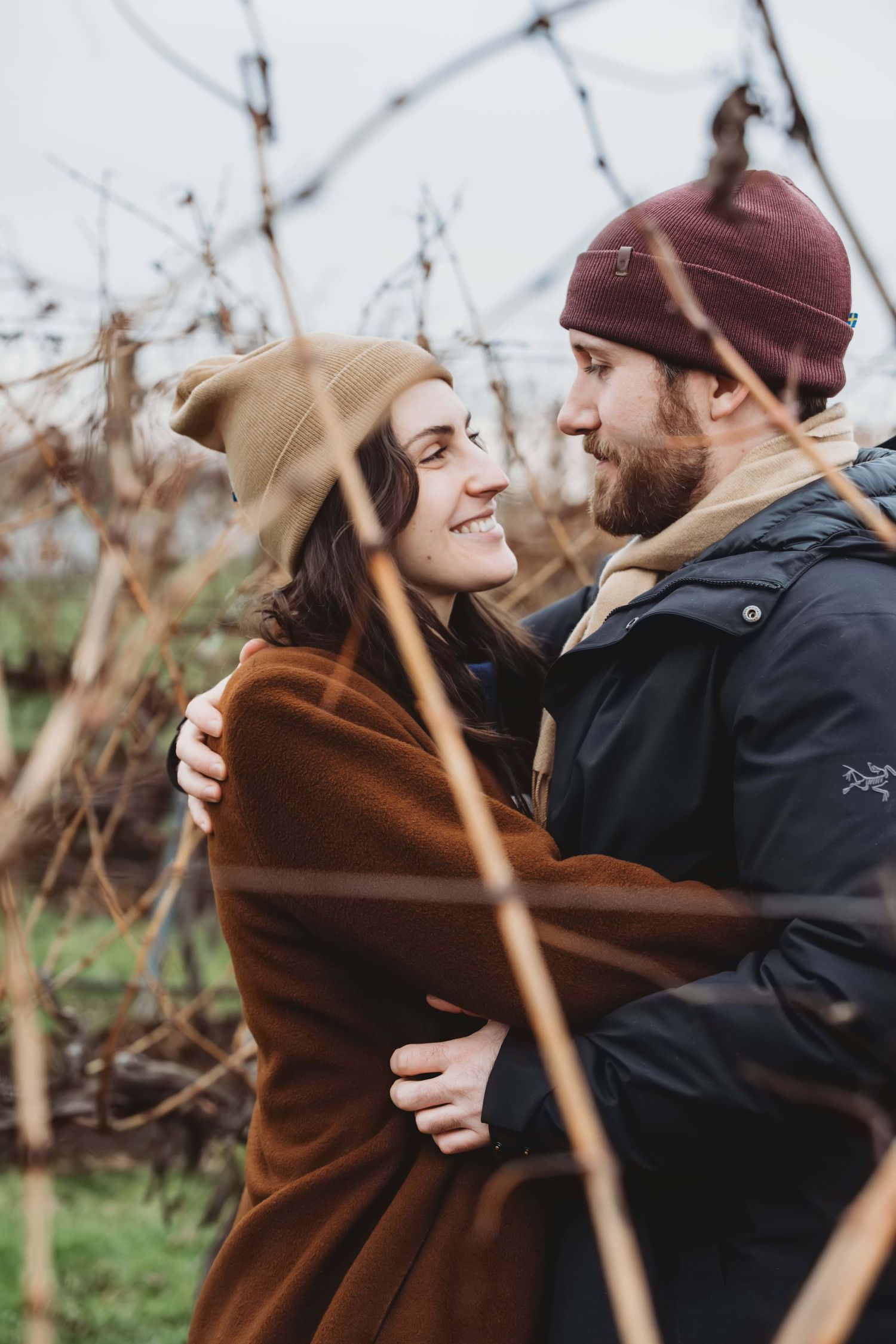 A couple looking at each other, photographed through brown dormant grape vines in the winter in Walla Walla.
