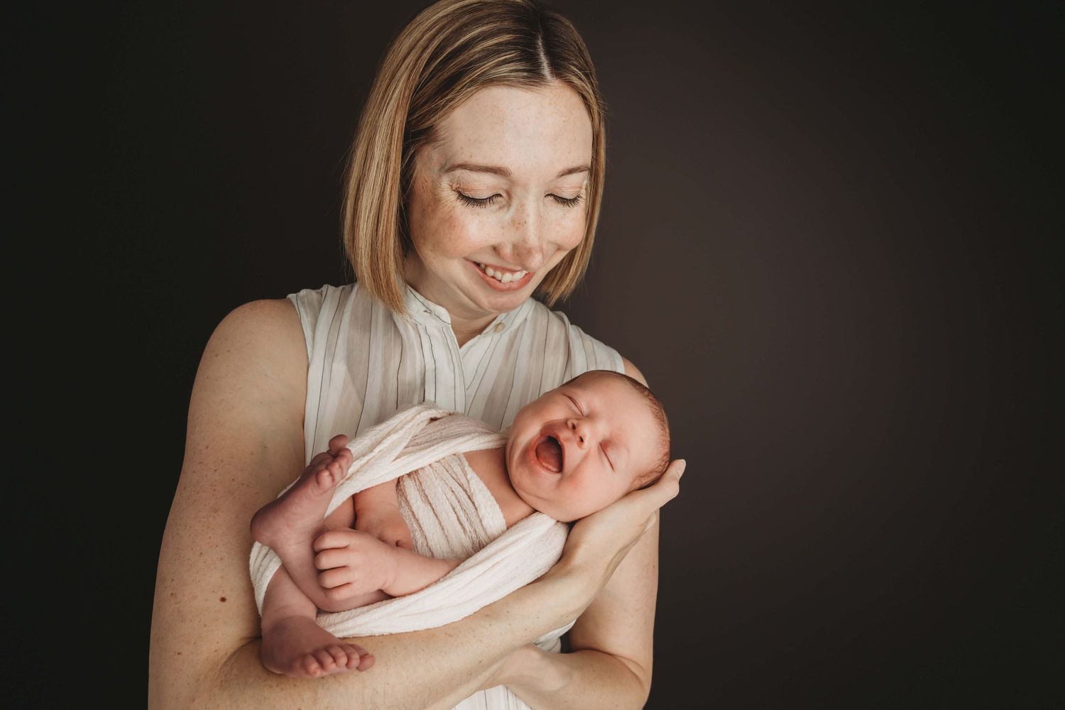 A mother looking down at her newborn as they both smile, on a black backdrop.