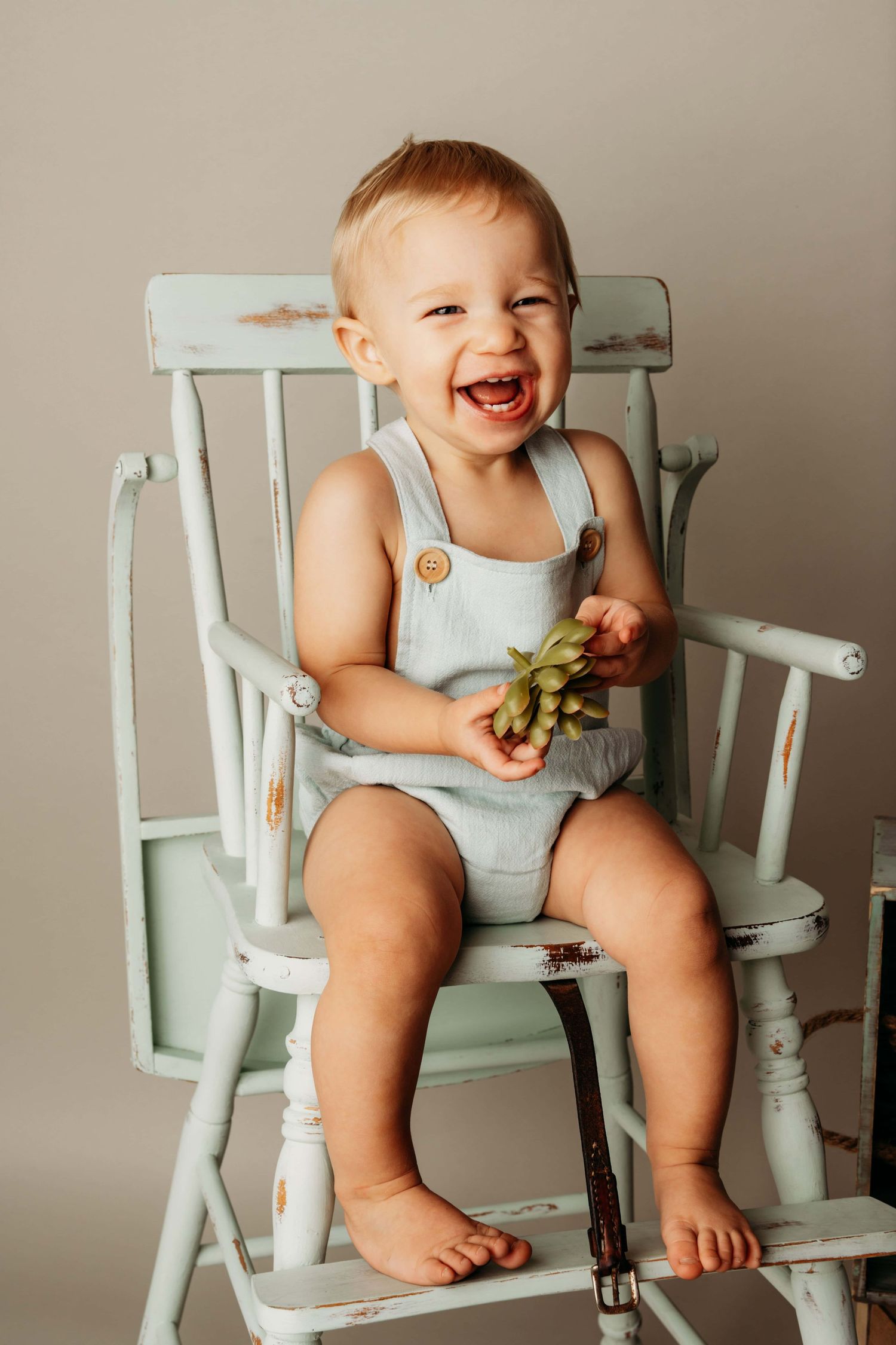 A one year old in a light blue romper sitting on an old wooden highchair holding a small succulent.