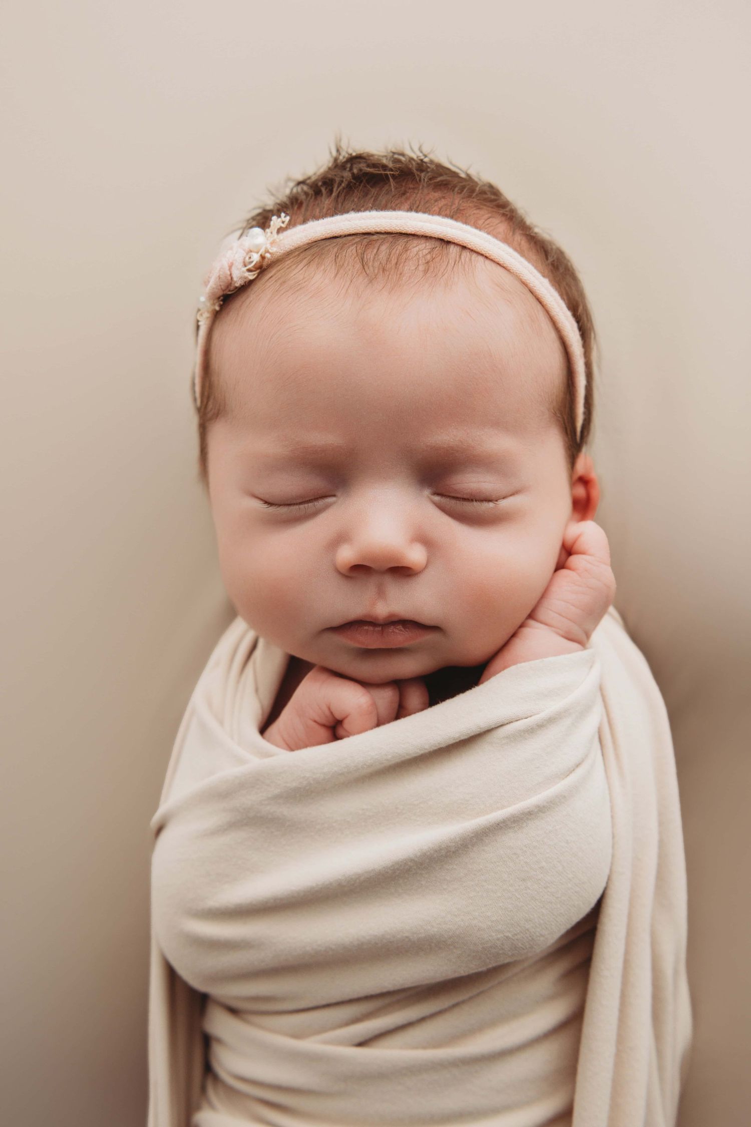 A newborn sleeping in neutral tones wearing a simple light pink headband.