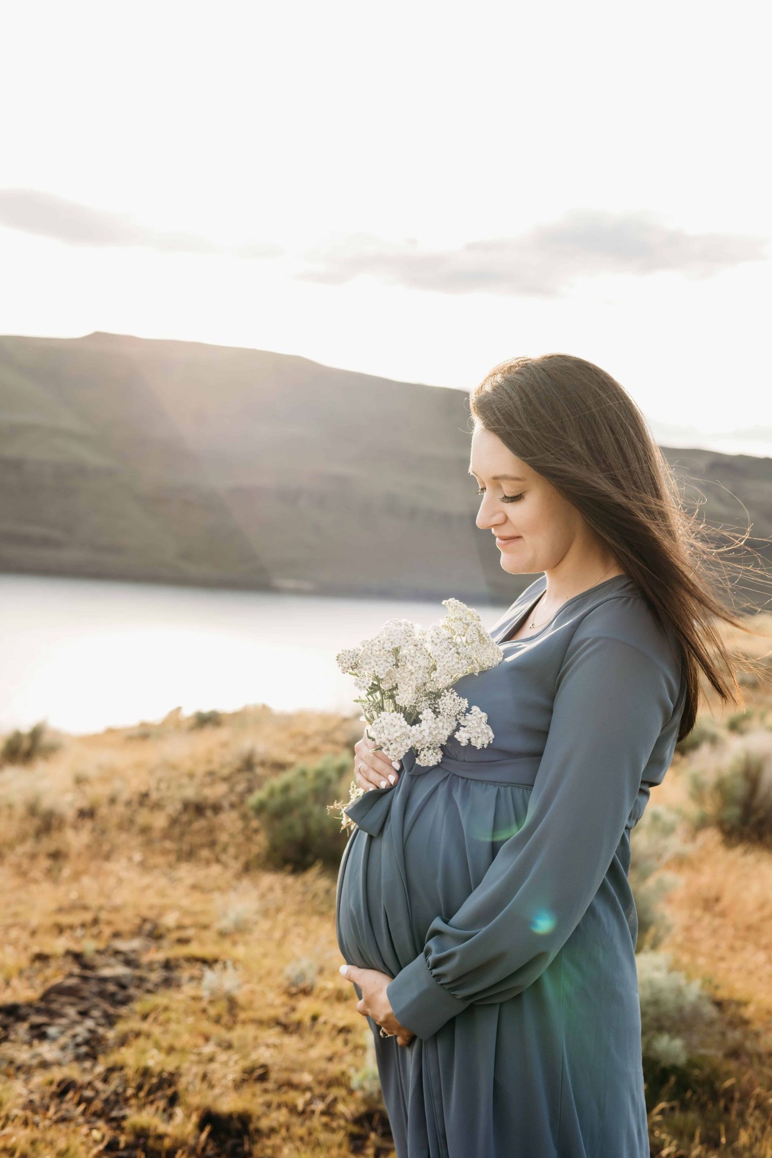 A portrait of an expecting mother wearing a dusty blue dress, holding white flowers in her hand and looking down at her belly.