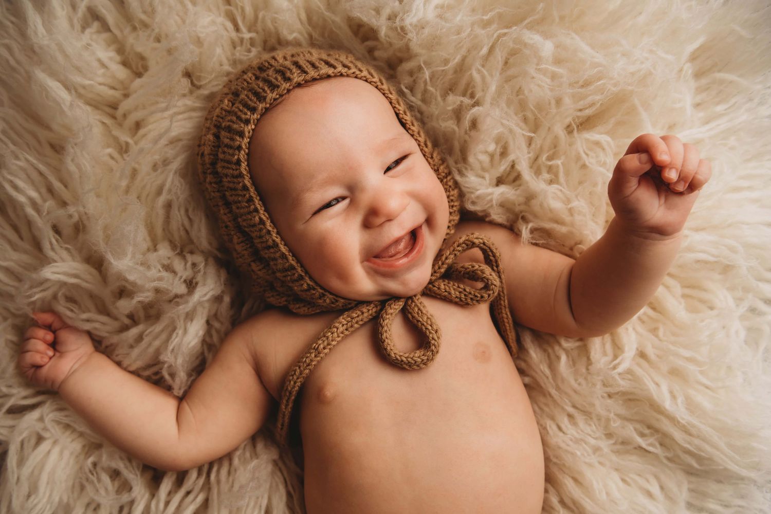 A portrait of a baby lying on a soft cream flokati wearing a brown hat and smiling big.