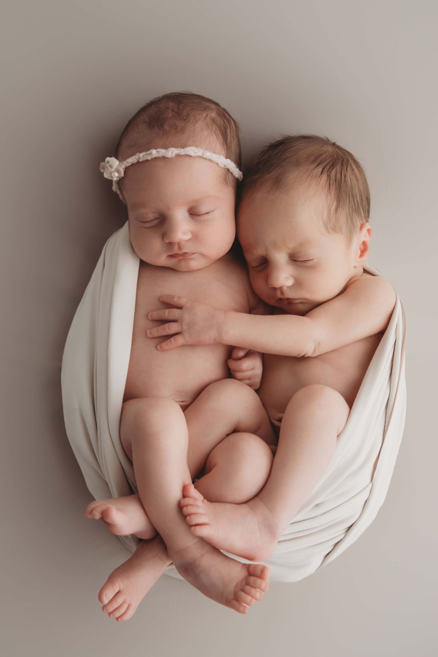 Boy and girl twins cuddle wrapped in a white blanket with their arms around each other.