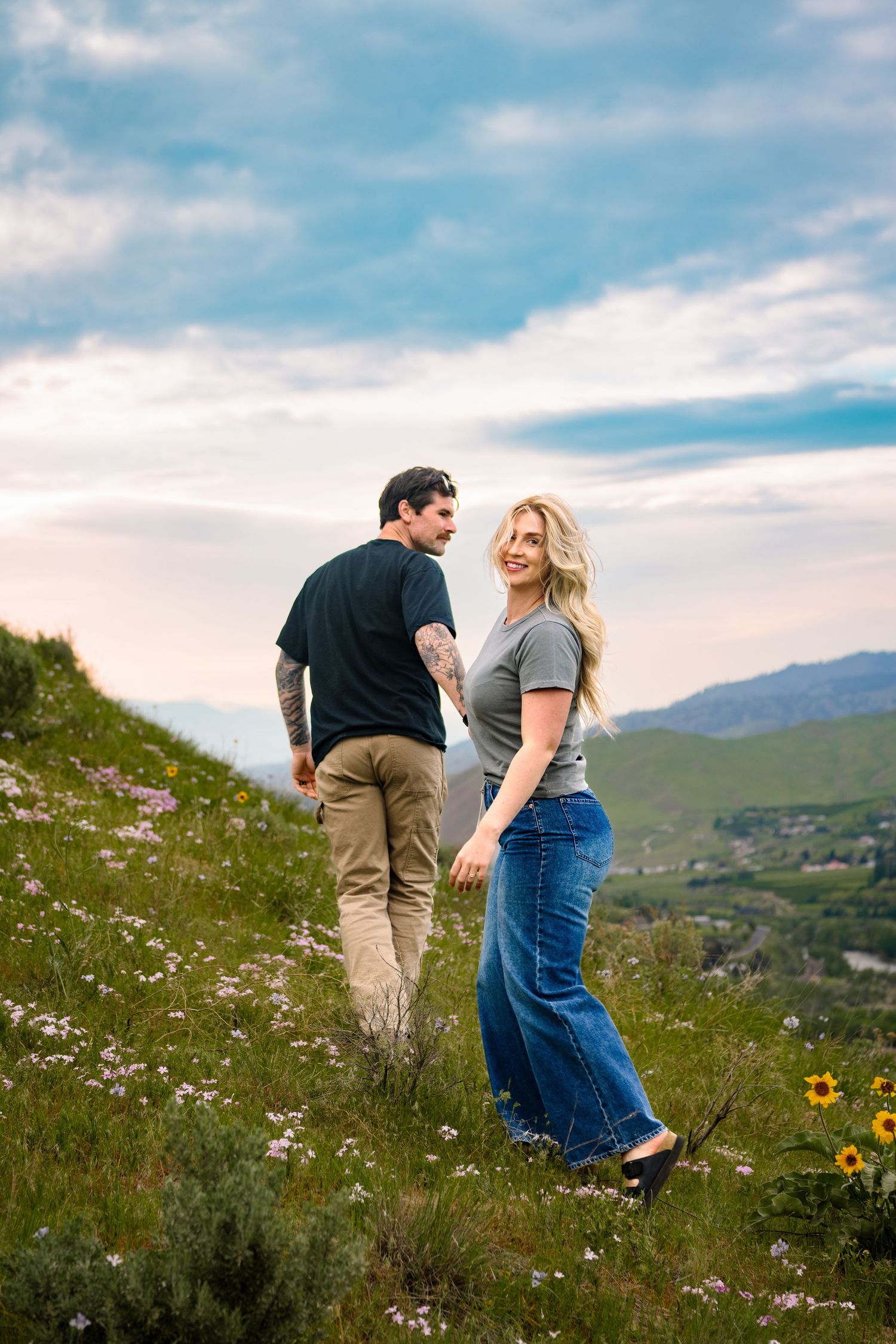 A couple walking through a wildflower field while the woman looks back at the camera.