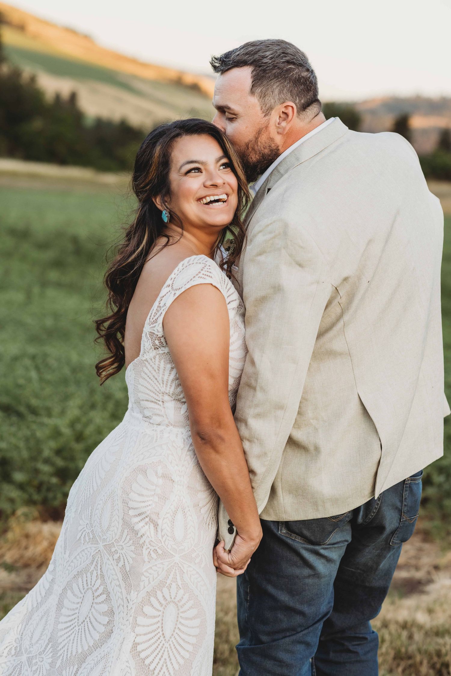 A bride laughing as her groom kisses the side of her head.