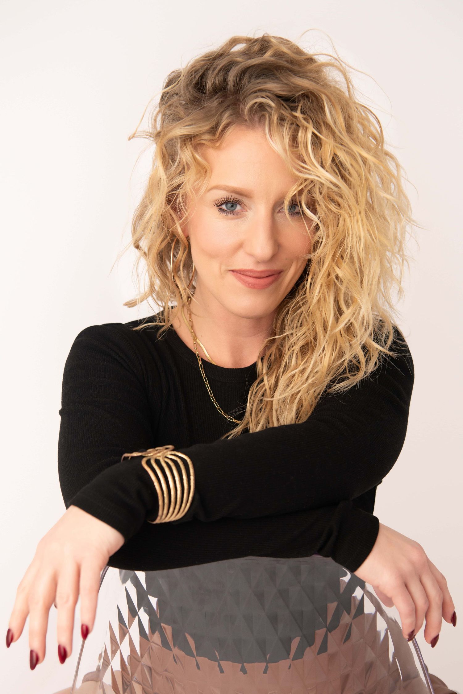 A professional headshot of a woman with curly blonde hair sitting on a clear acrylic chair.