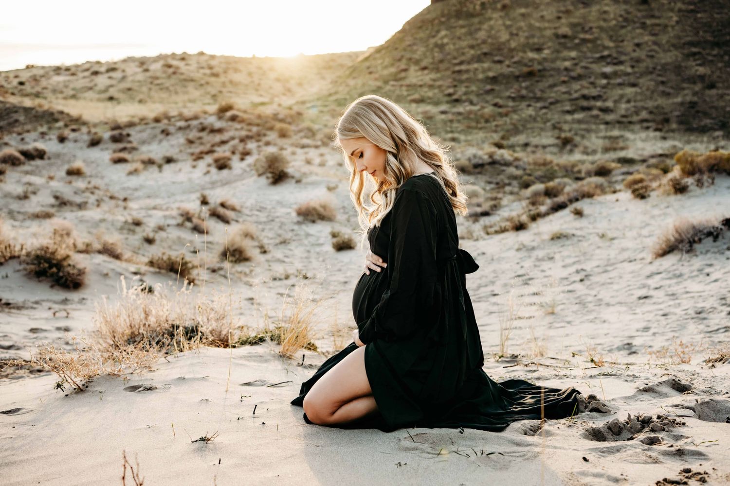 A pregnant woman sitting in sand, wearing a black dress and looking down at her belly.