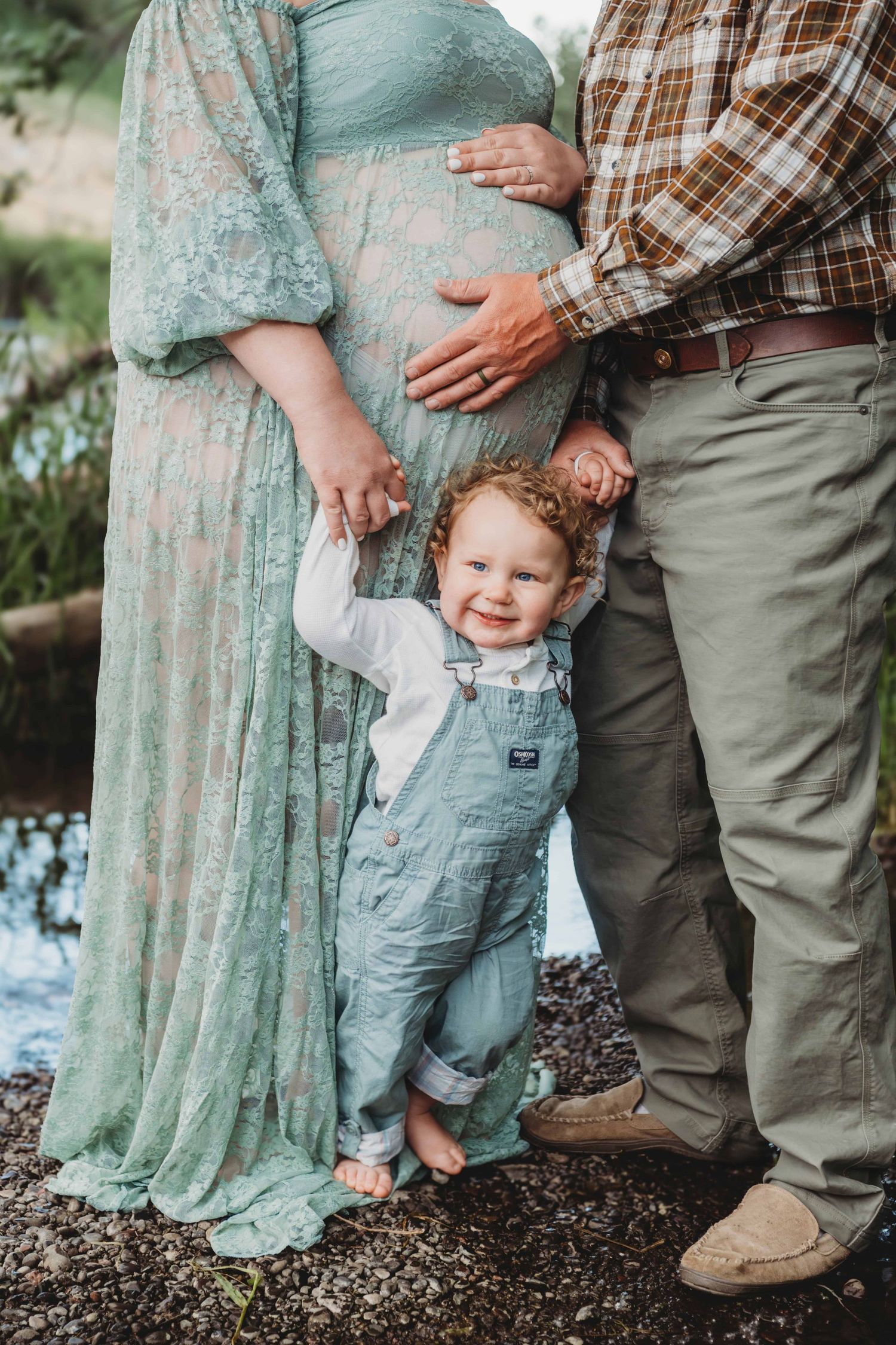 A toddler stands near his parents while they place their hands on the baby bump during a maternity session with a Walla Walla photographer.