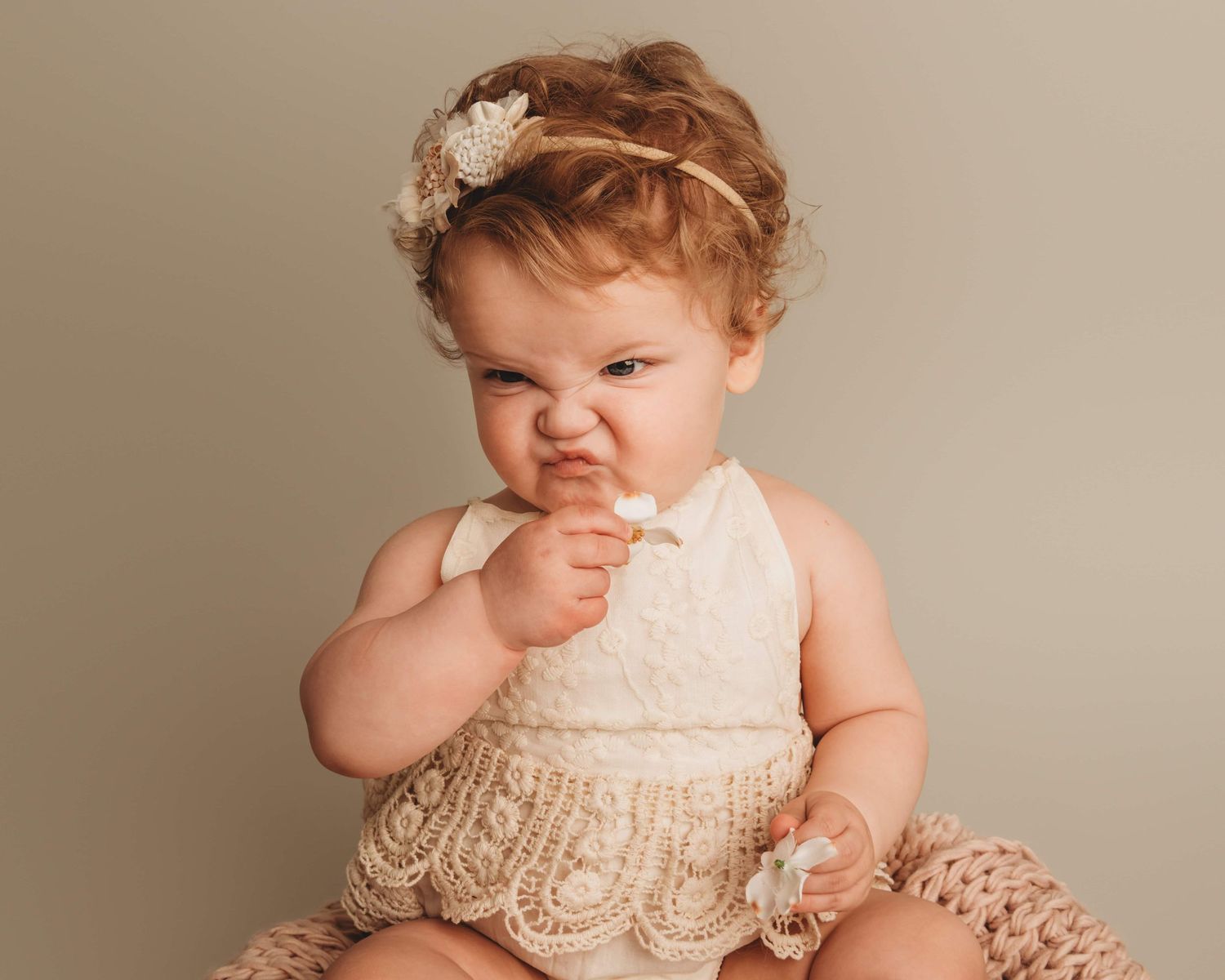A baby with curly brown hair sniffing a flower and making a funny face.