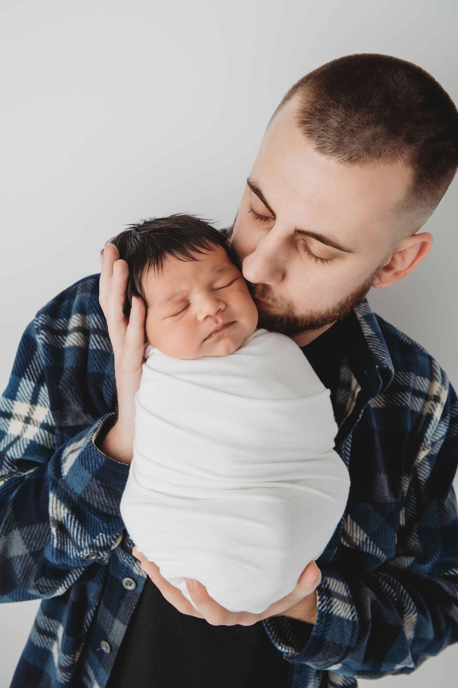 A dad kissing his newborn babies cheek on a white backdrop.