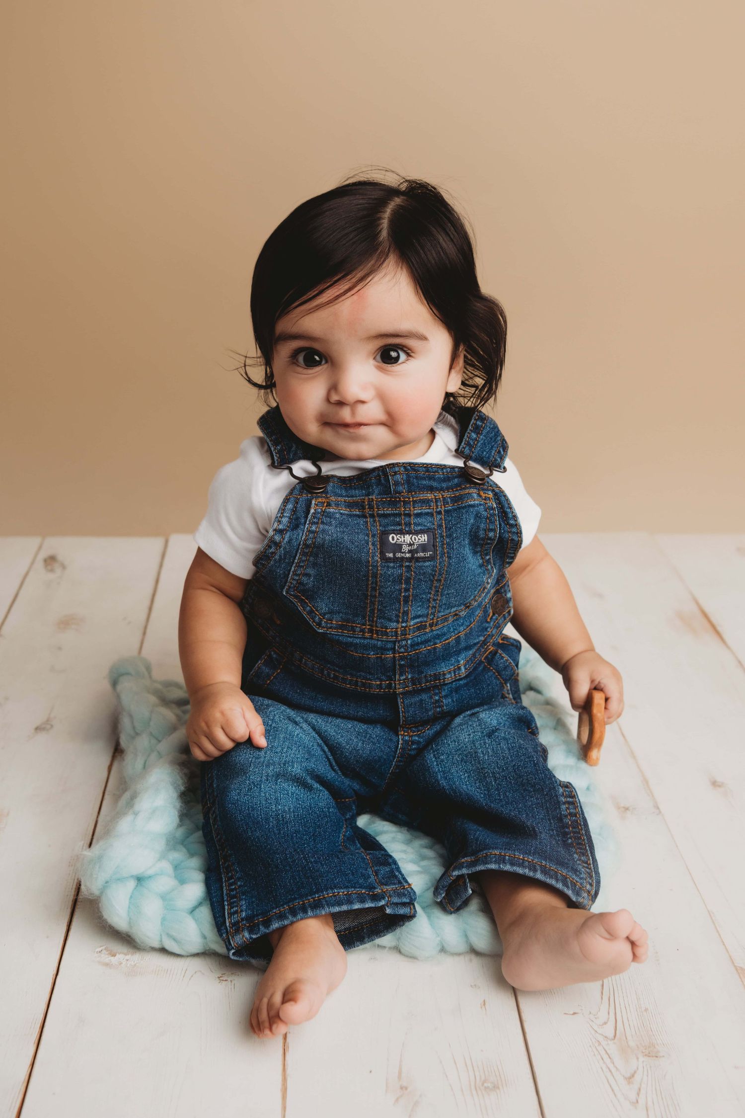 A baby wearing jean overalls sitting on a blue knitted mat with a soft smile.