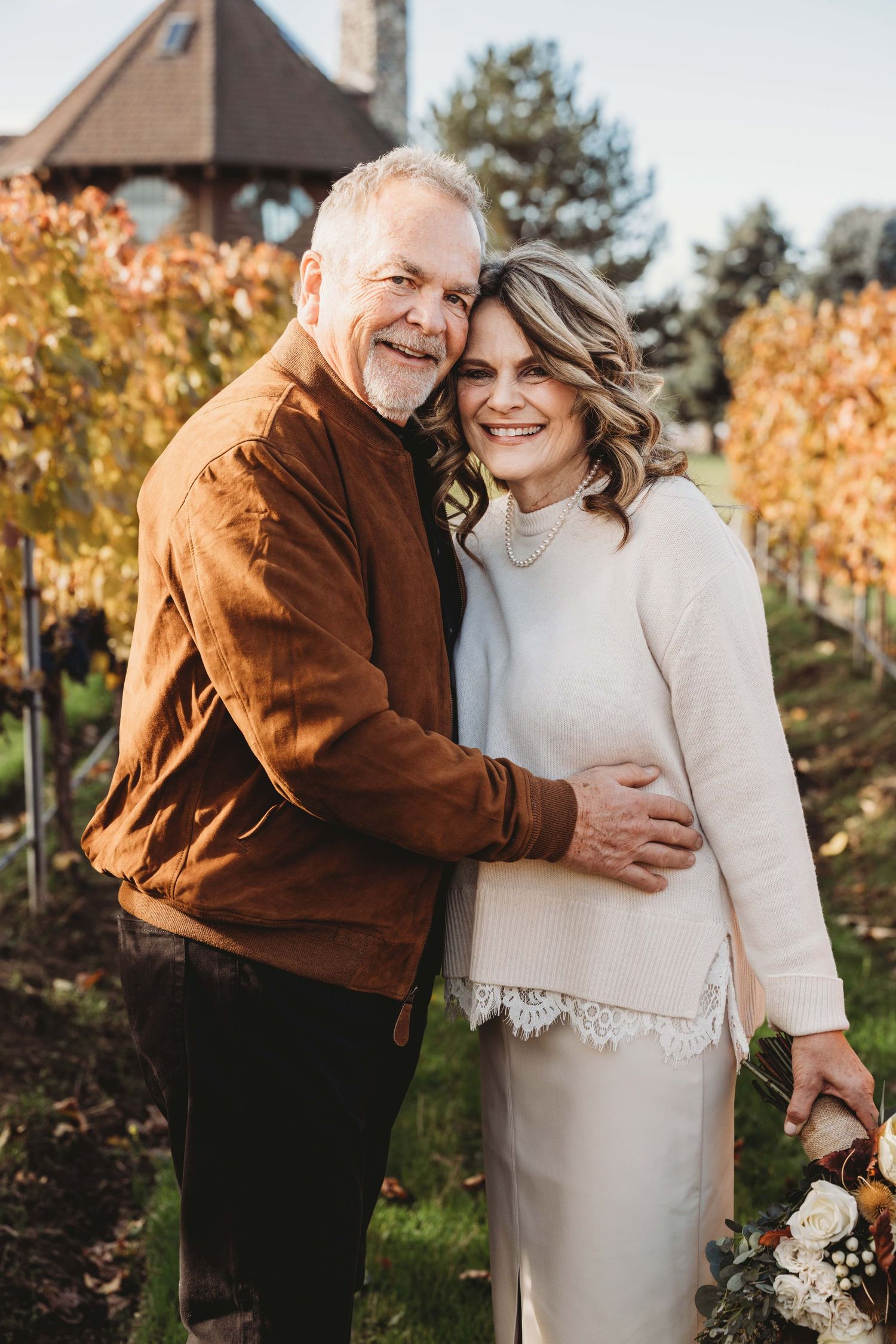 A couple standing in a vineyard with fall colors and smiling at the camera.