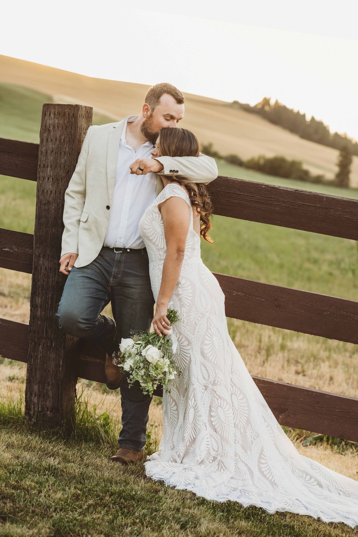 A groom leaning against a wooden fence with his arm around his bride.