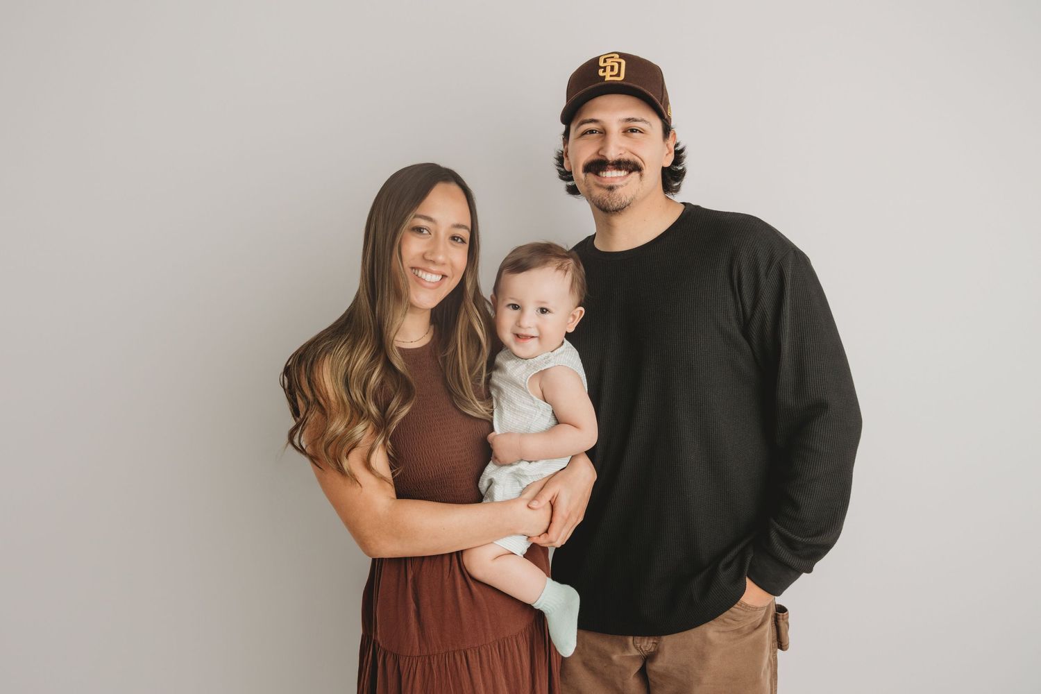 A family of three with their one year old baby smiling at the camera on a white backdrop.