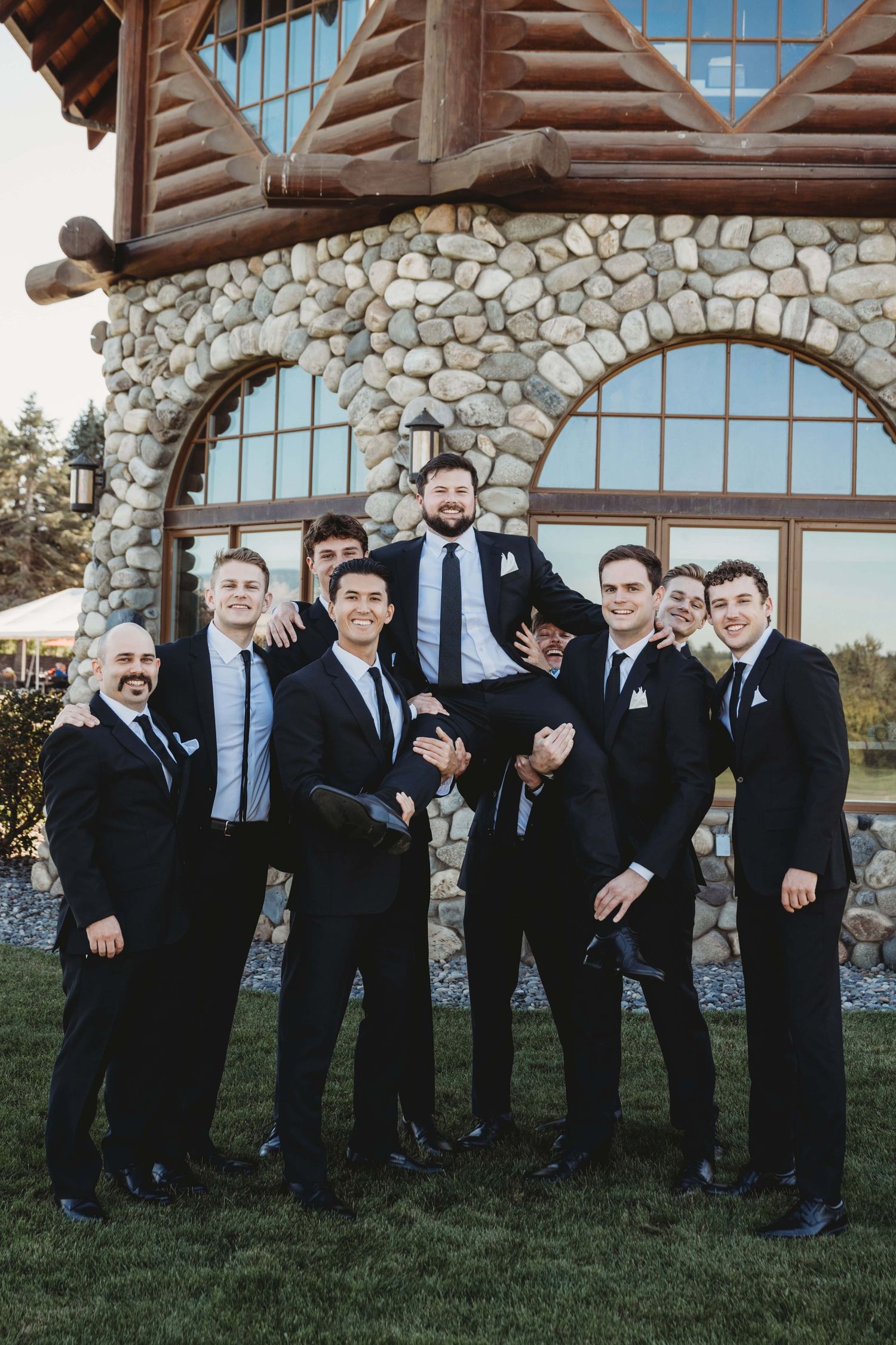 A group of groomsmen wearing black suits picking up the groom.