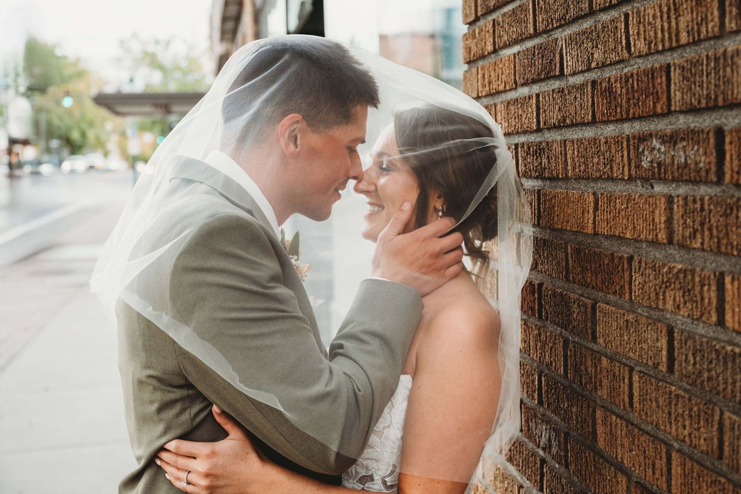 A bride and groom under a veil leaning against a brick building.