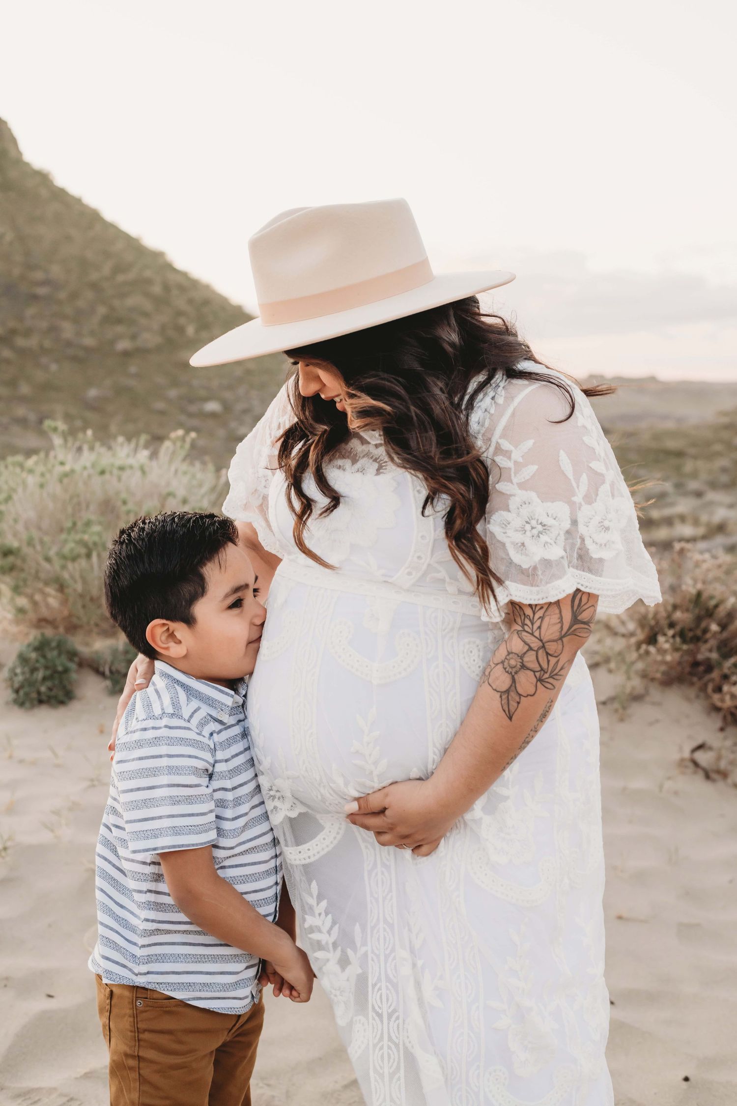 A boy kissing his mom's pregnant belly in a desert scene.