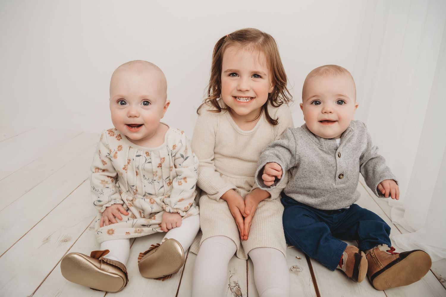 A girl and her twin baby siblings sit on a white wooden floor and smile for a classic portrait at a Walla Walla photography studio.