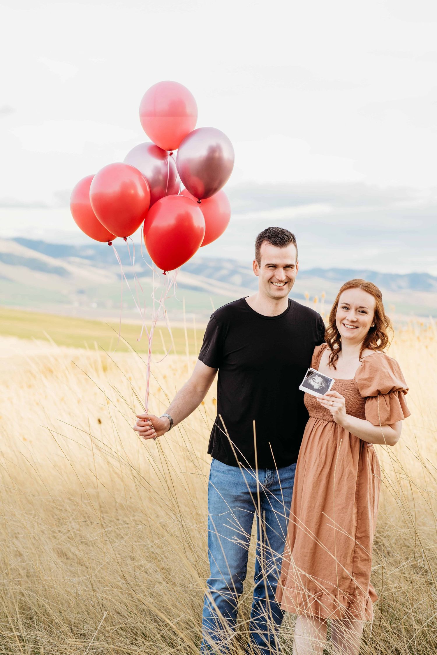 A couple holding an ultrasound photo and pink balloons to announce that they are expecting a baby girl.